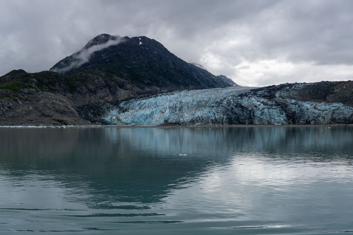 A massive tidewater glacier meets the sea beneath cloudy skies in an Alaskan fjord.