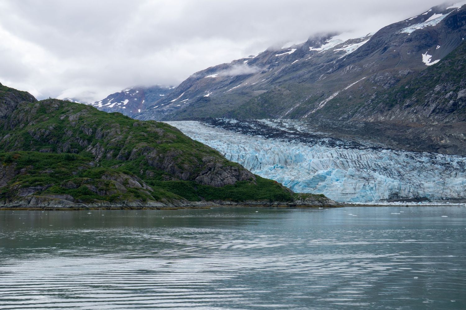 Blue glacier descends between green mountains into calm waters under overcast skies.