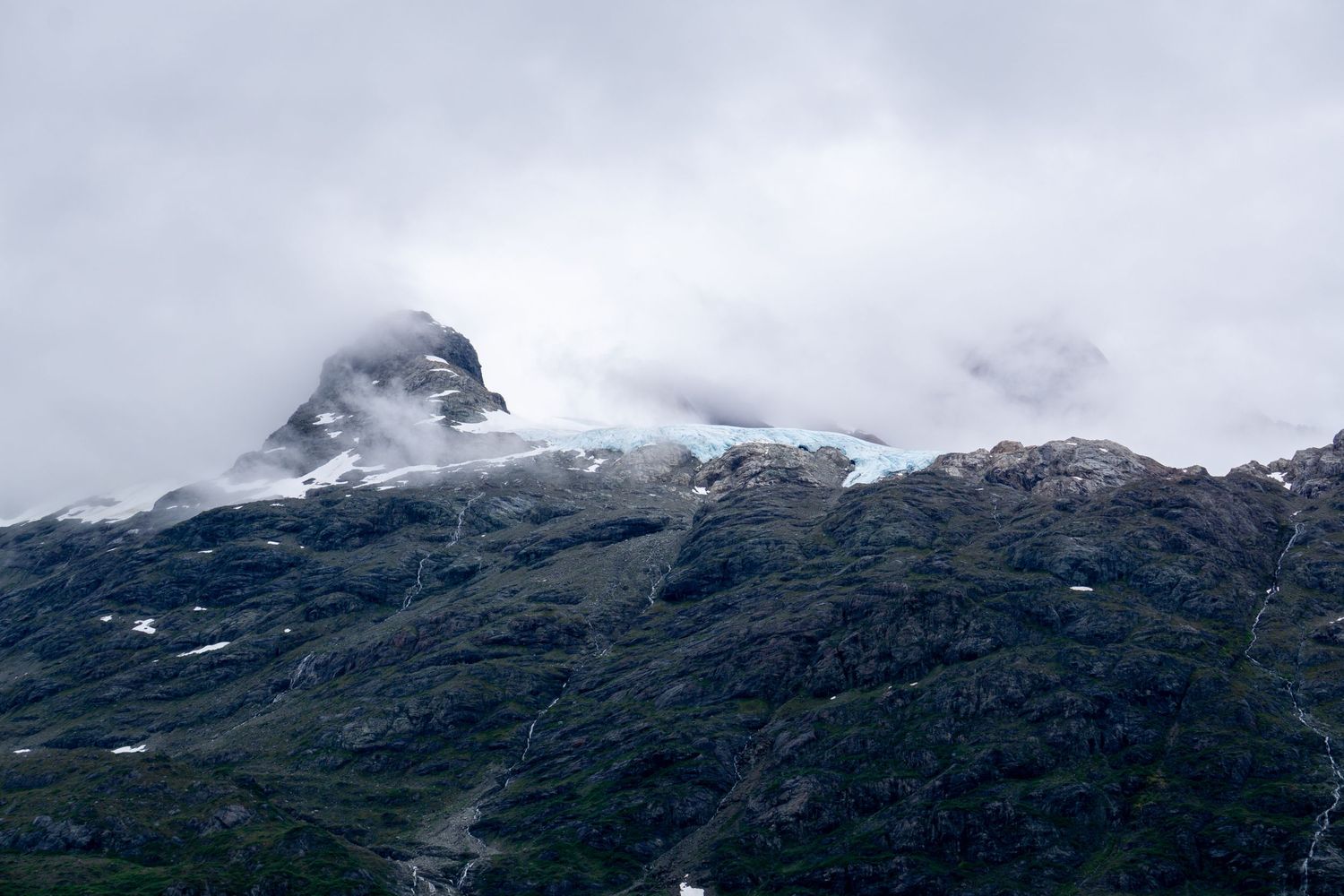 Snow-capped mountain peak emerges through dense cloud cover in rugged Alaskan terrain.