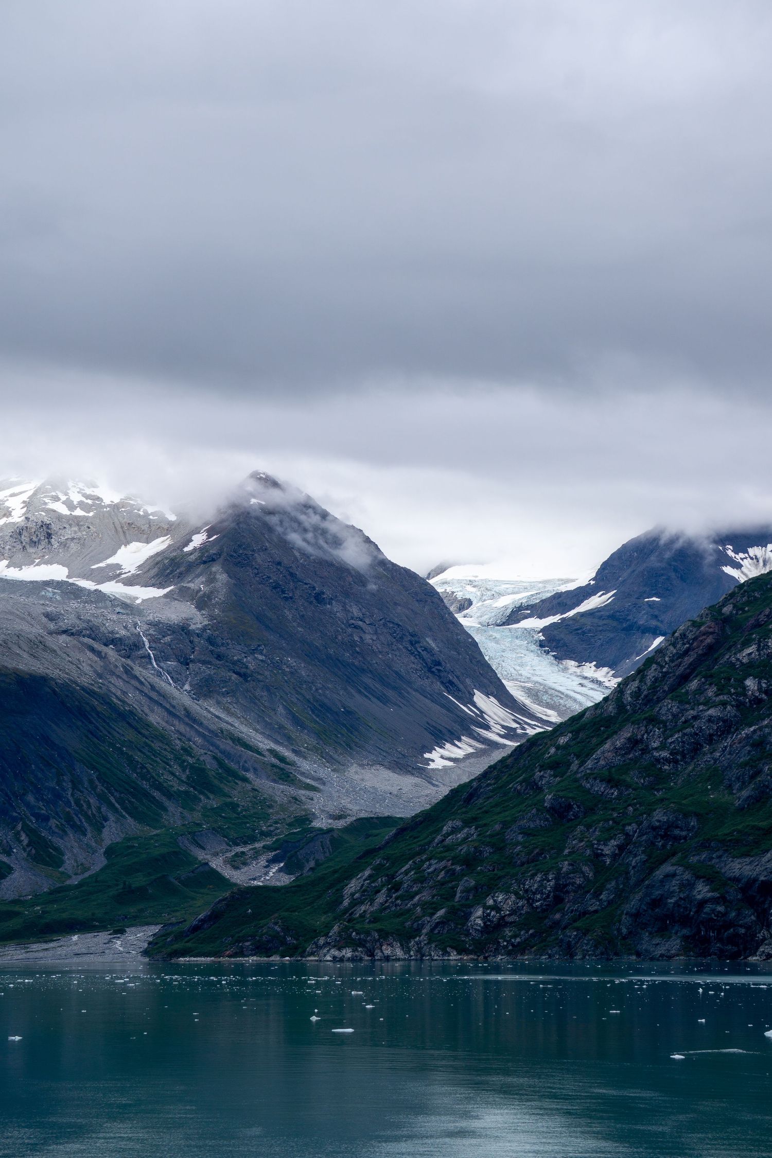 Glacier flows through mountain valley into turquoise fjord waters under moody sky.