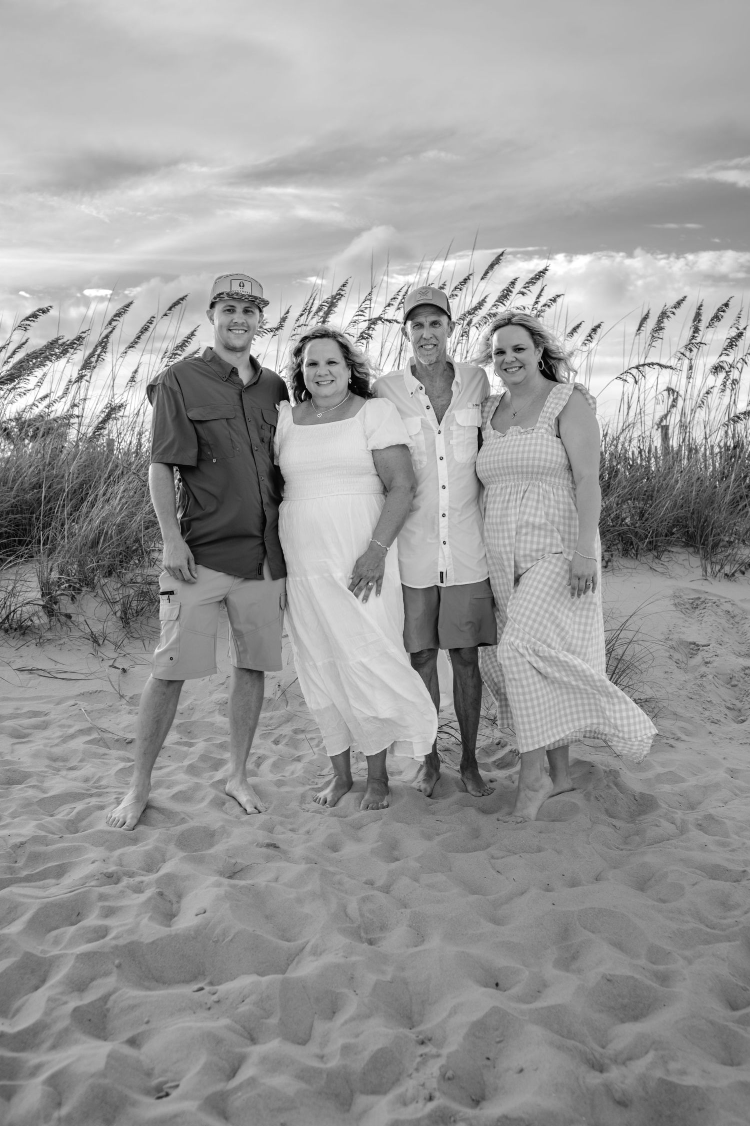 Black and white photo of four people smiling together on a sandy beach with sea oats and dramatic clouds behind them.