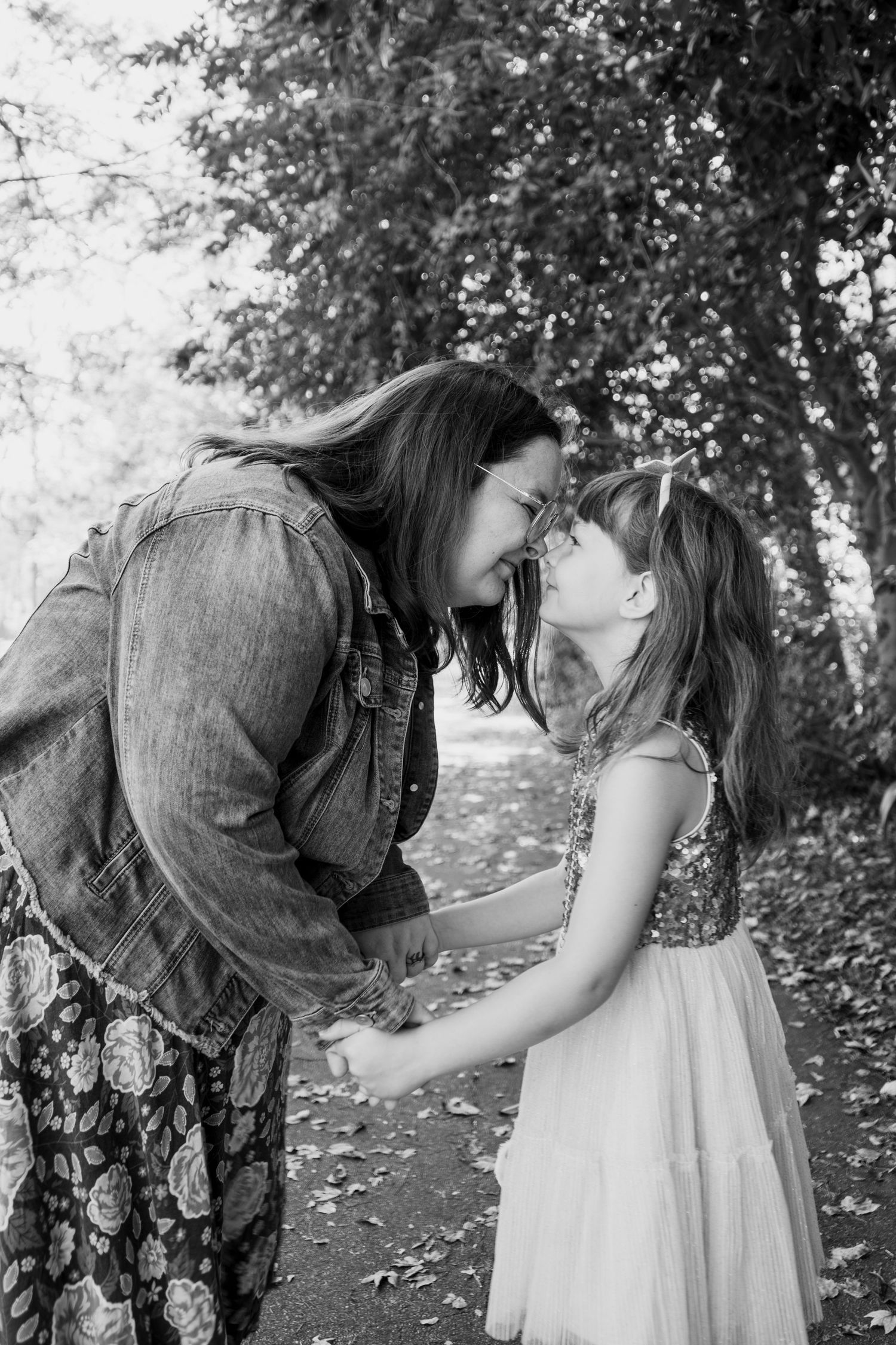 Black and white photo of a mother and young daughter touching foreheads and holding hands on a tree-lined path.