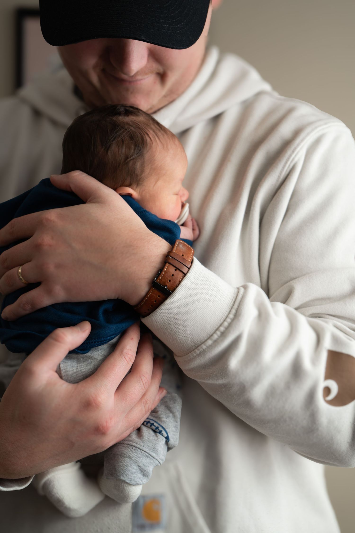 Smiling father in a white Carhartt hoodie and cap gently cradling his newborn baby in a blue outfit.