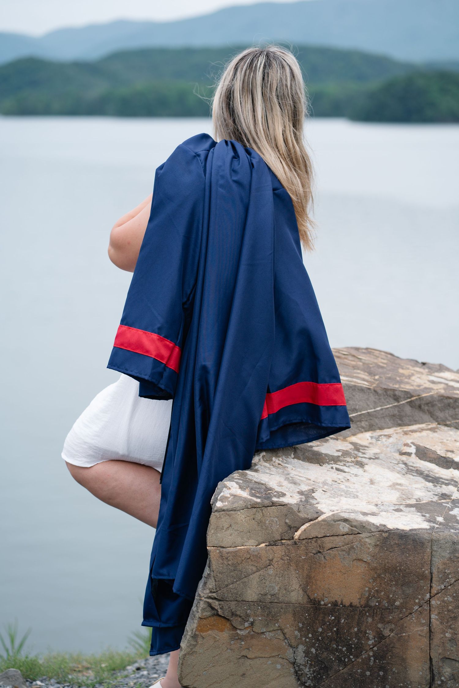 Blonde woman in white dress holding navy graduation gown sitting on rocks overlooking a lake.