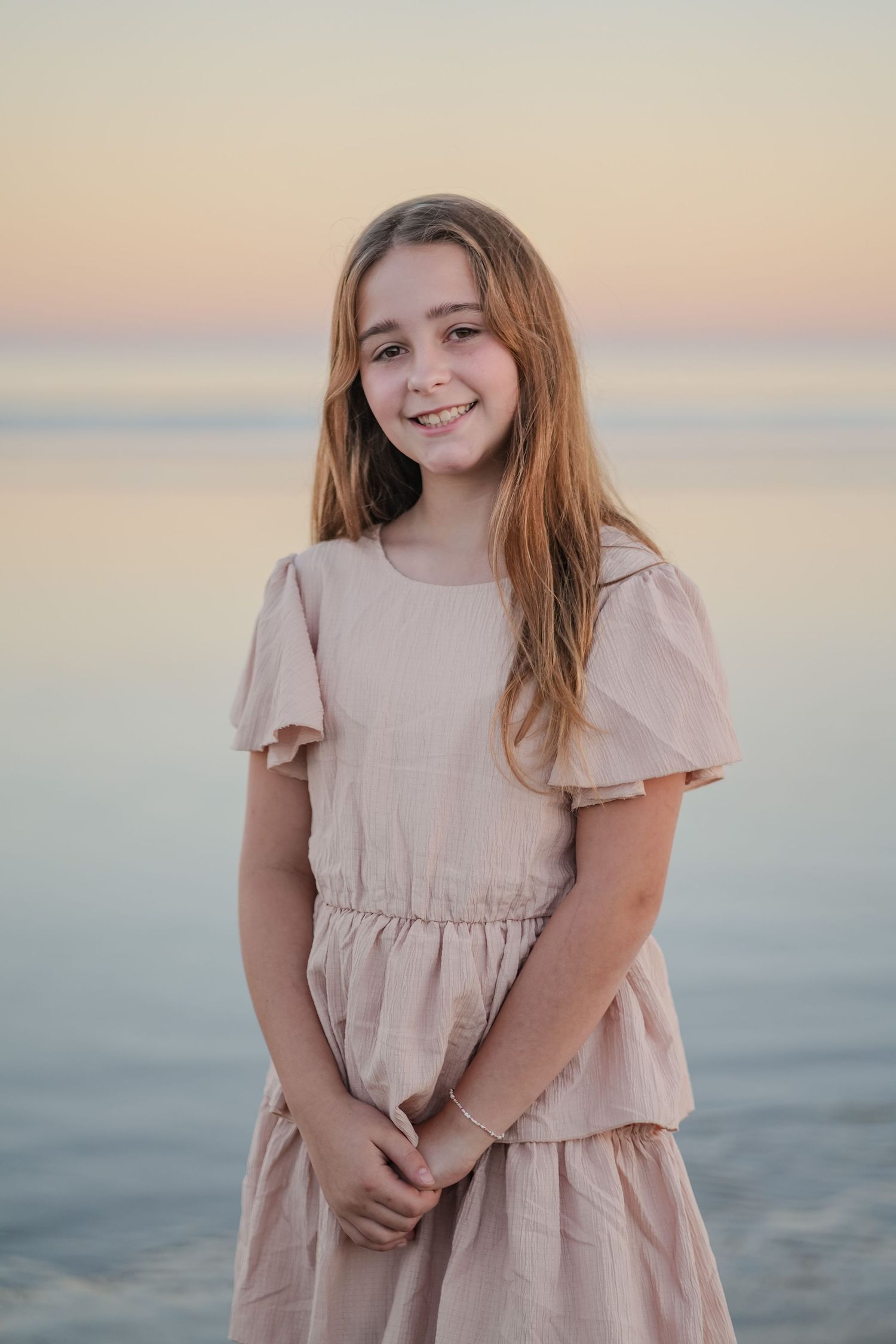 Happy preteen girl in a blush pink outfit smiling on a beach at golden hour.