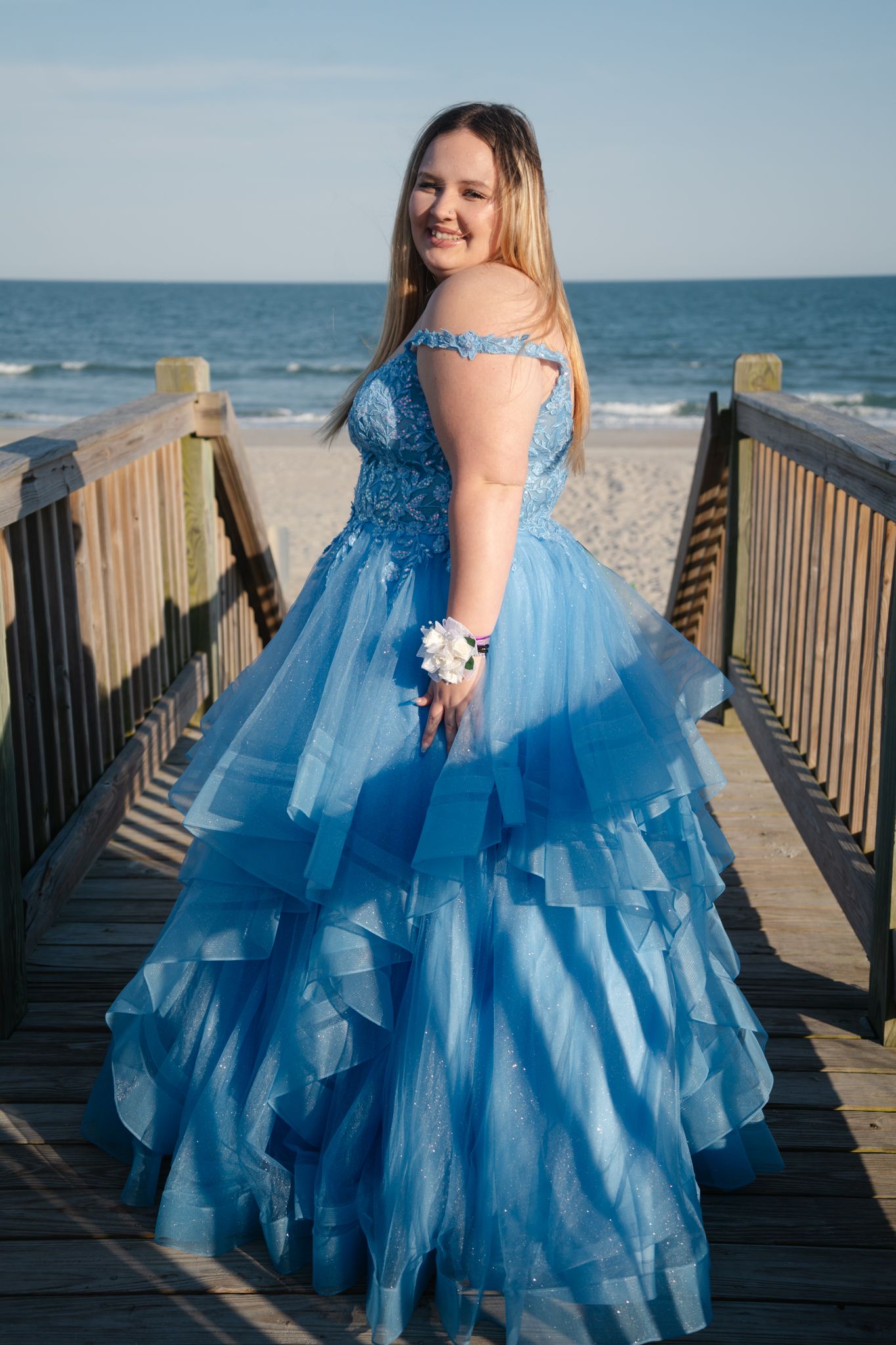 Young woman in layered blue prom gown with wrist corsage posing on beach boardwalk at sunset.