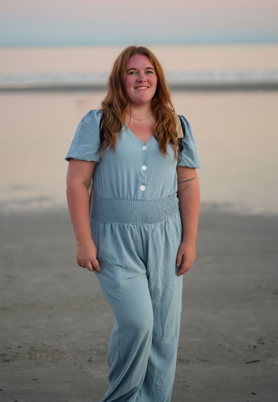 Smiling redhead woman in light blue jumpsuit walking on beach at sunset.