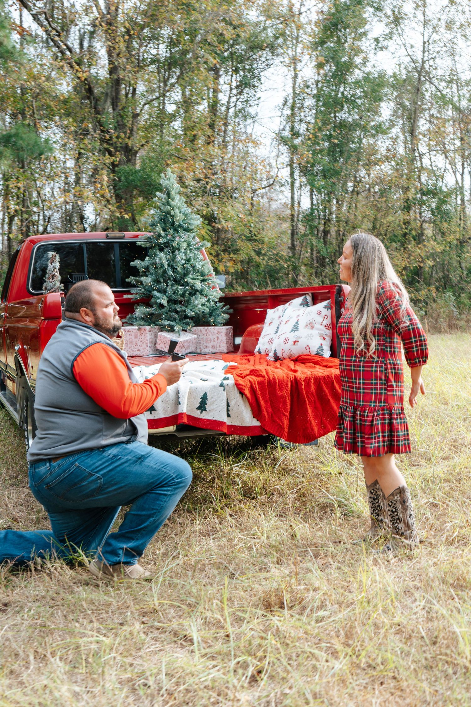 Couple enjoying Christmas gifts at a red truck bed decorated with holiday pillows and a tree in a wooded setting.