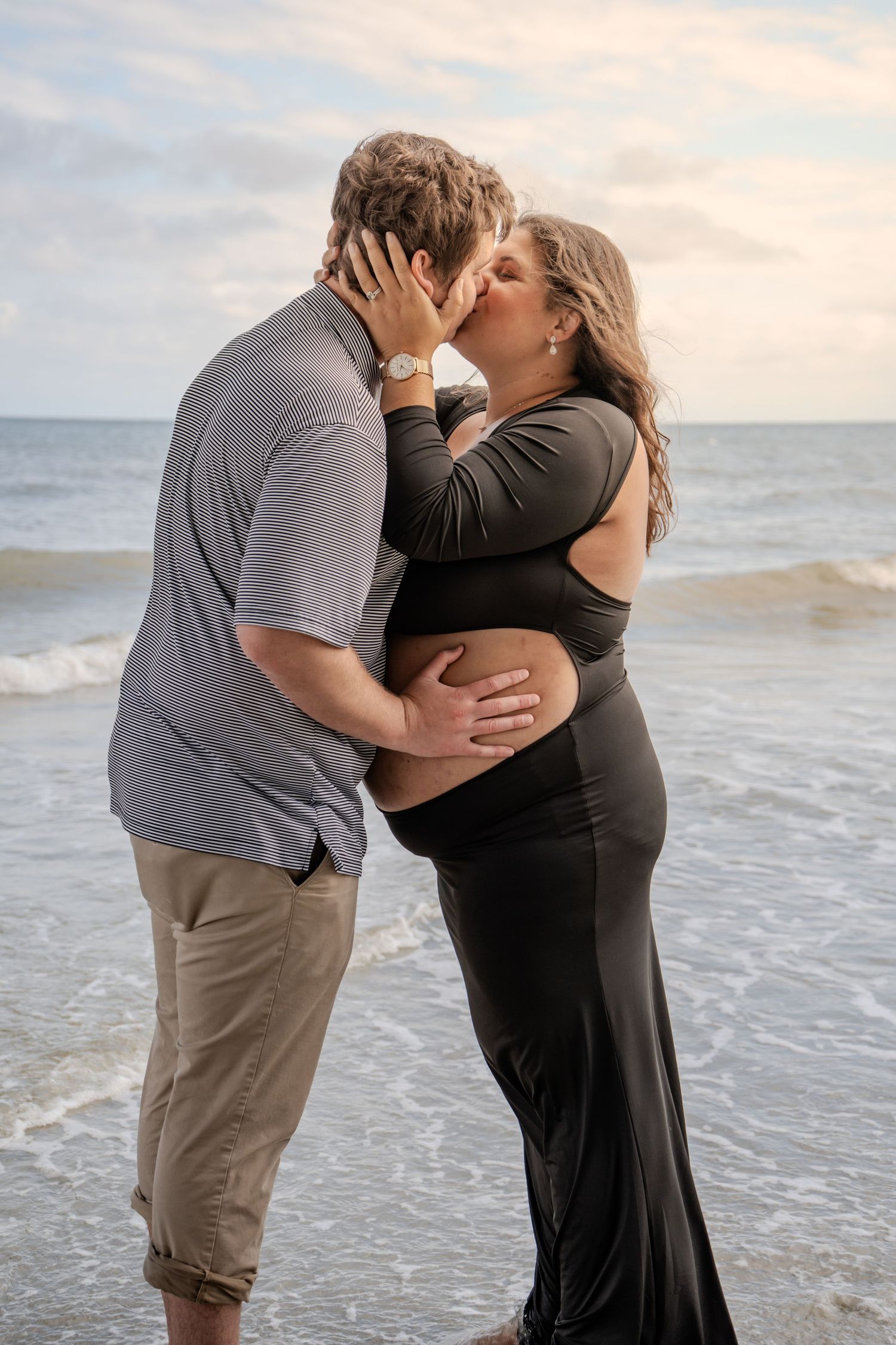 Couple sharing a romantic kiss on the beach during a maternity photoshoot, with ocean waves in the background.
