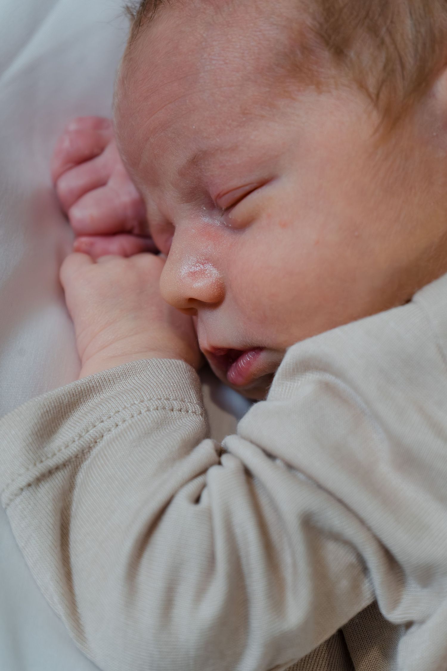 A sleeping newborn baby in a beige onesie with tiny fist raised near face.