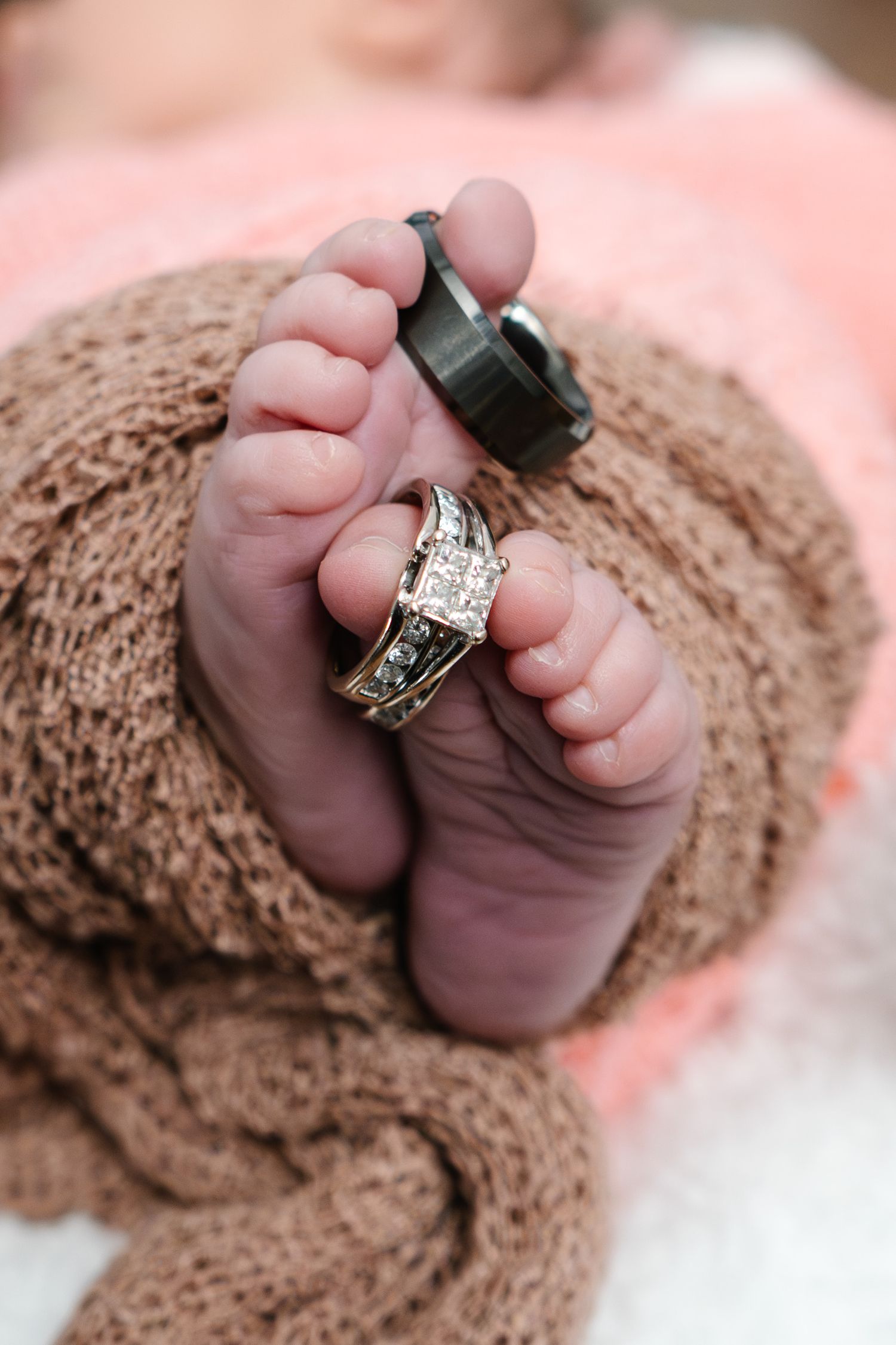 Close-up of newborn baby feet wrapped in brown knit blanket with diamond engagement ring and black band on tiny toes.