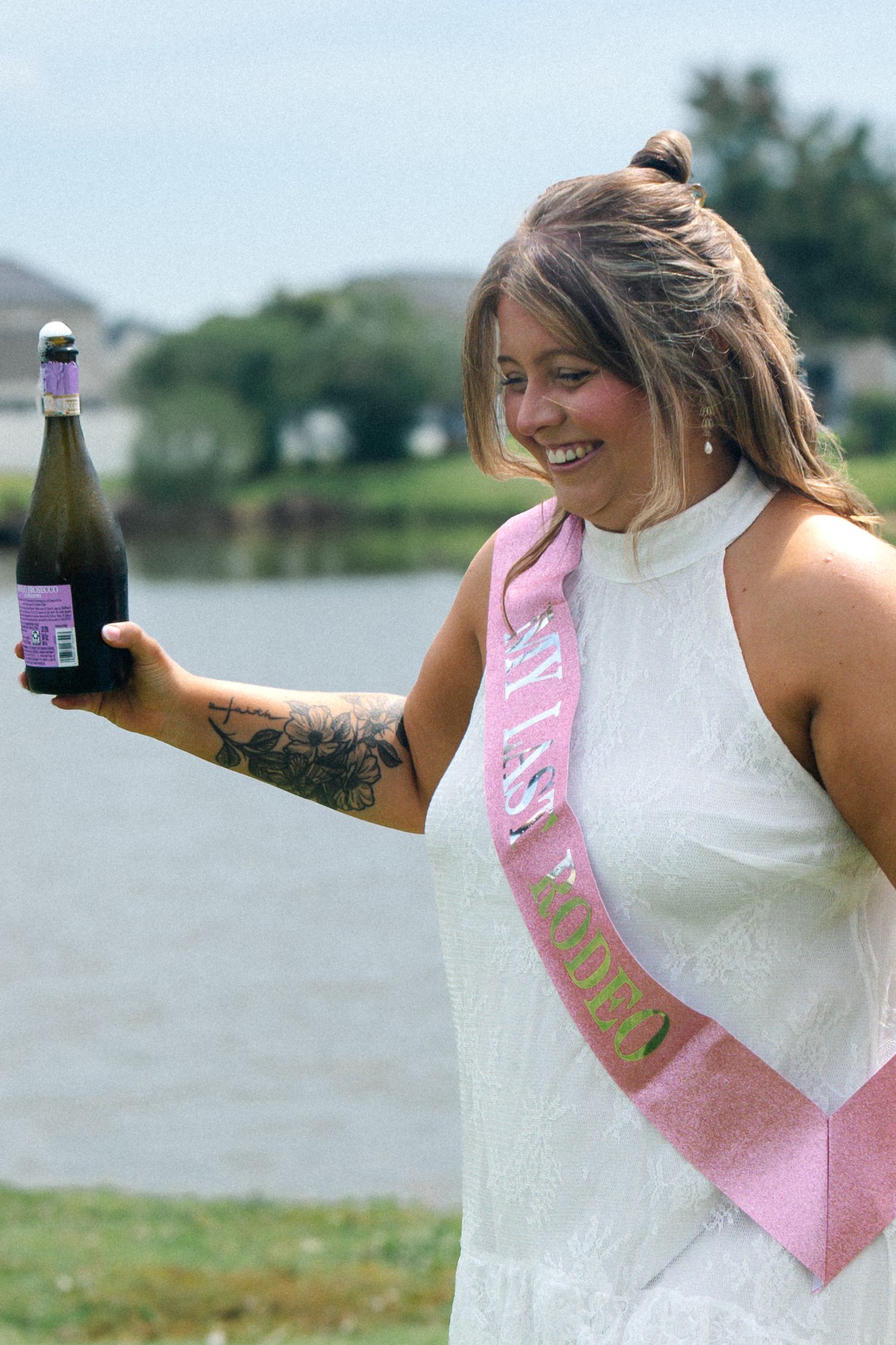 Woman in white dress wearing pink sash holds champagne bottle outdoors near lake, smiling at celebration event.
