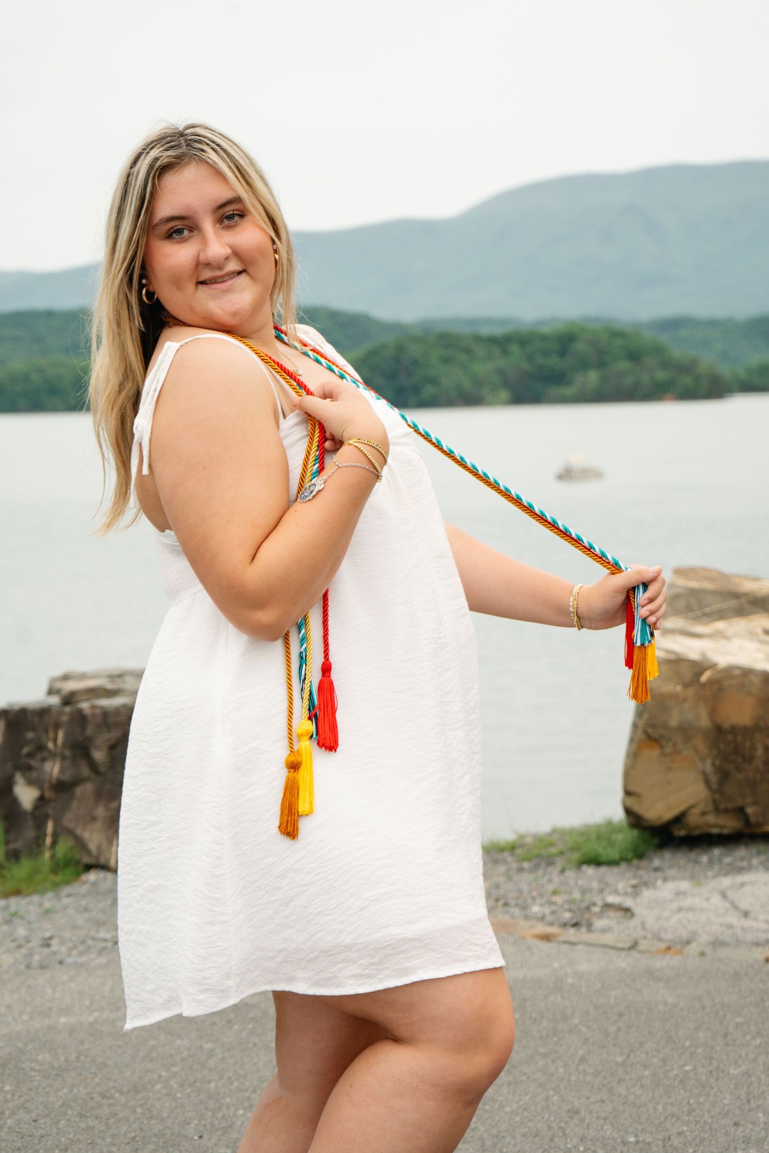 Smiling blonde woman in white dress holding colorful graduation honor cords near scenic lake.