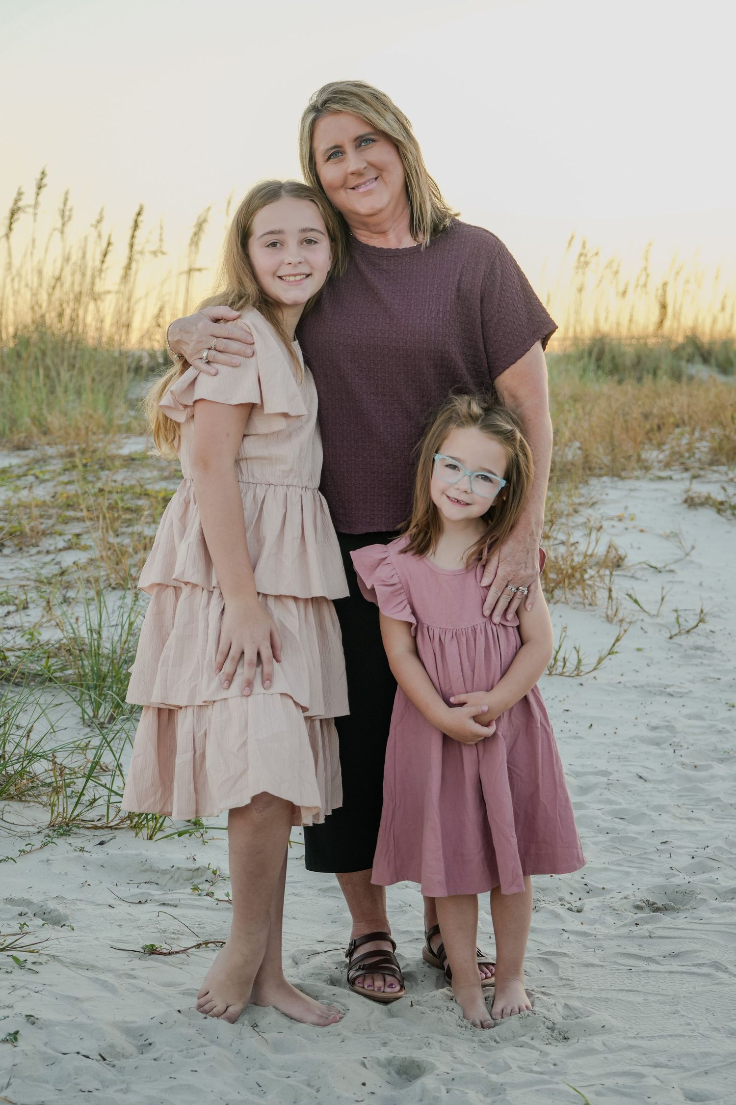 A mother and her two daughters in coordinating pink and mauve dresses posing on a sandy beach.