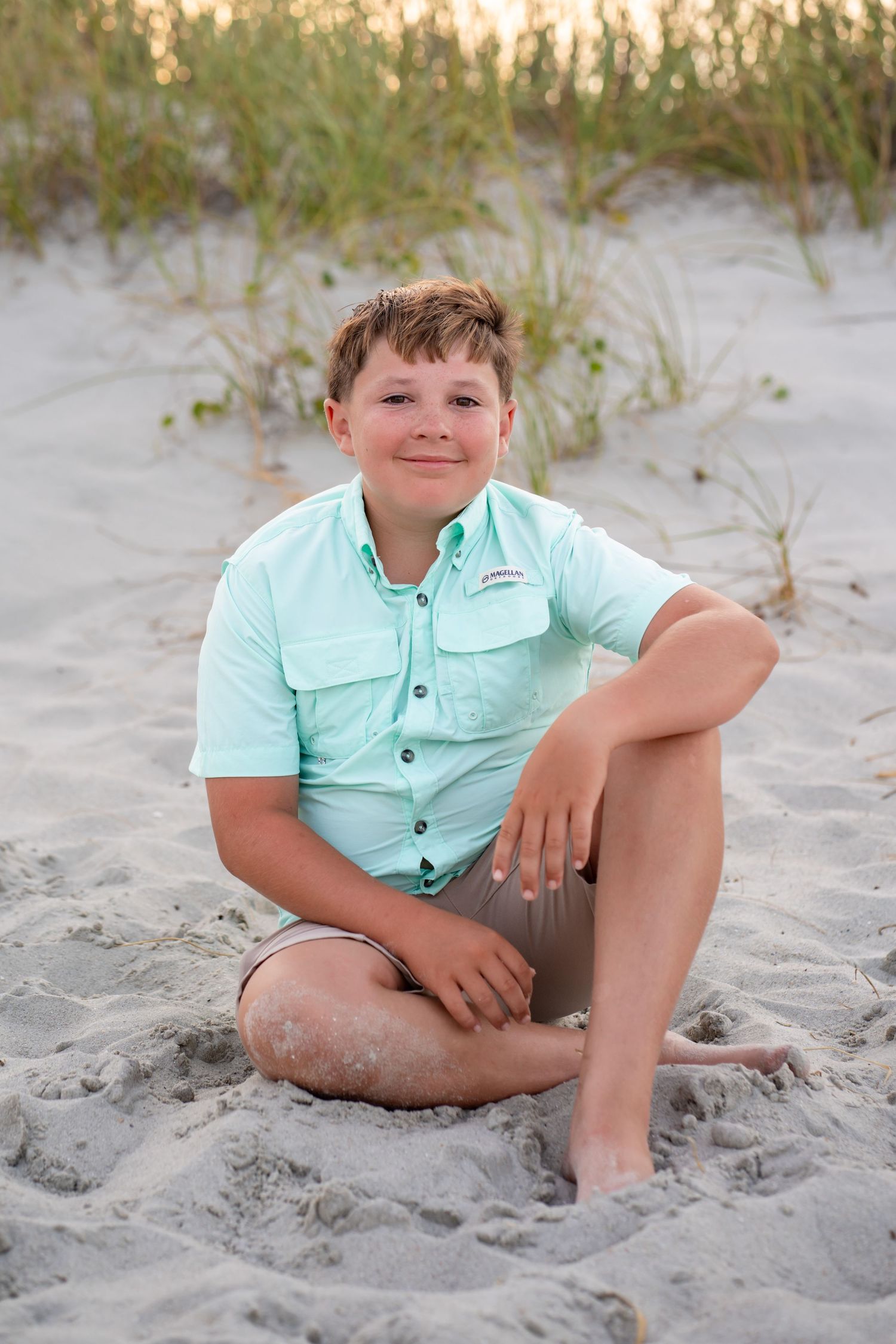 A smiling boy in a mint green shirt sits cross-legged on sandy beach dunes with sea grass in the background.