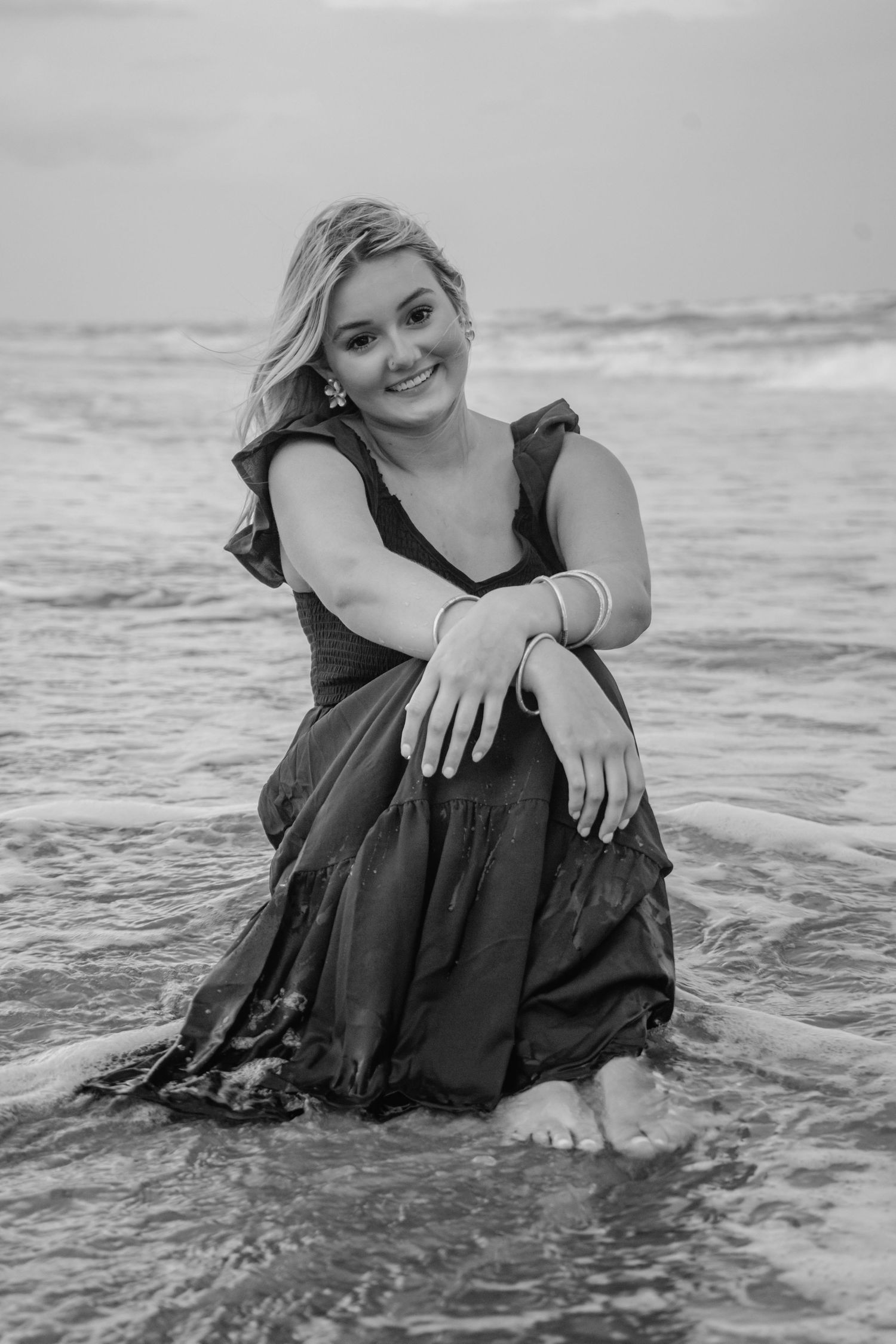 Black and white photo of a smiling blonde woman in a dress sitting in ocean waves on a beach.