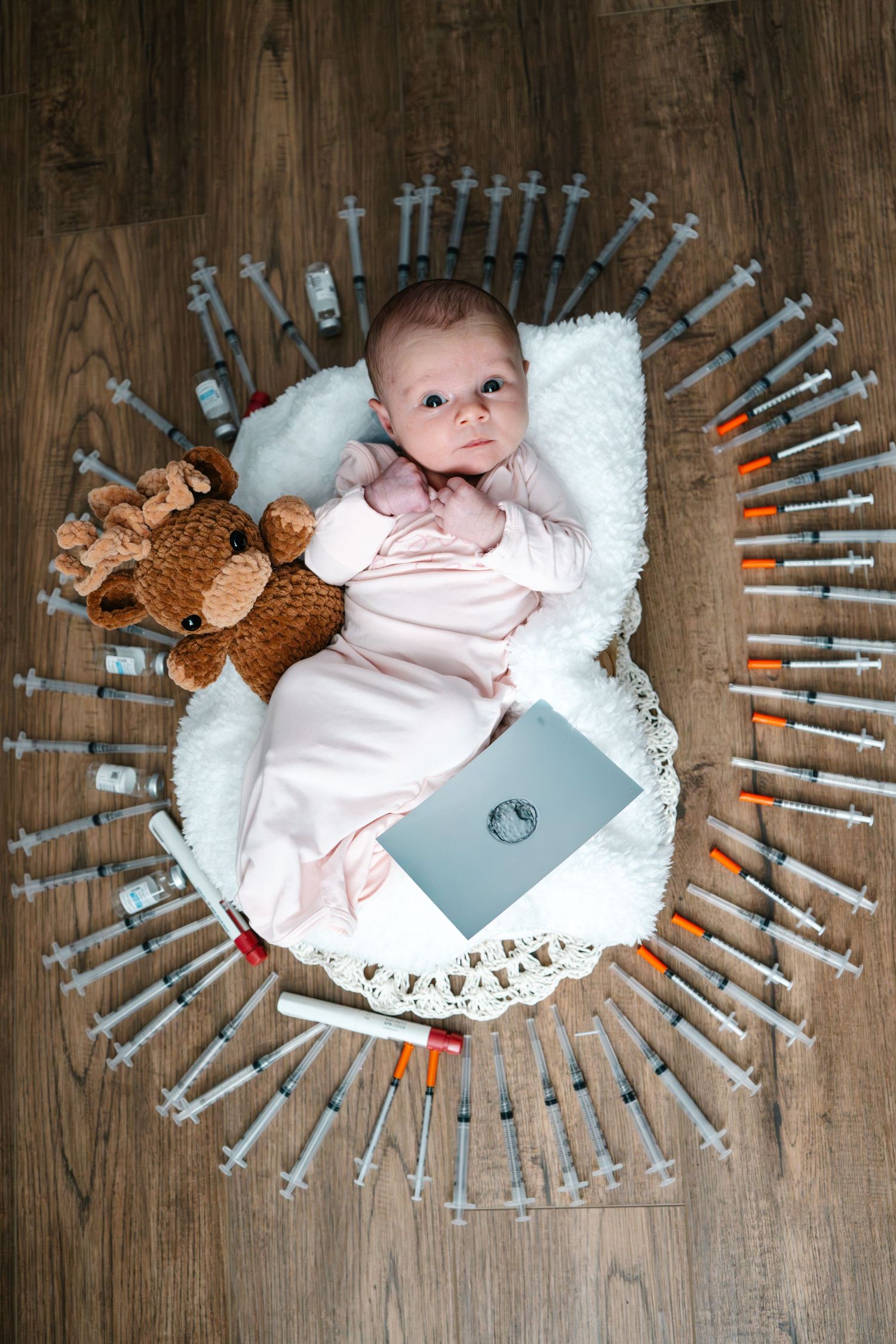 IVF photo, Newborn baby in pink onesie lying in white basket with teddy bear, surrounded by syringes on wooden floor.