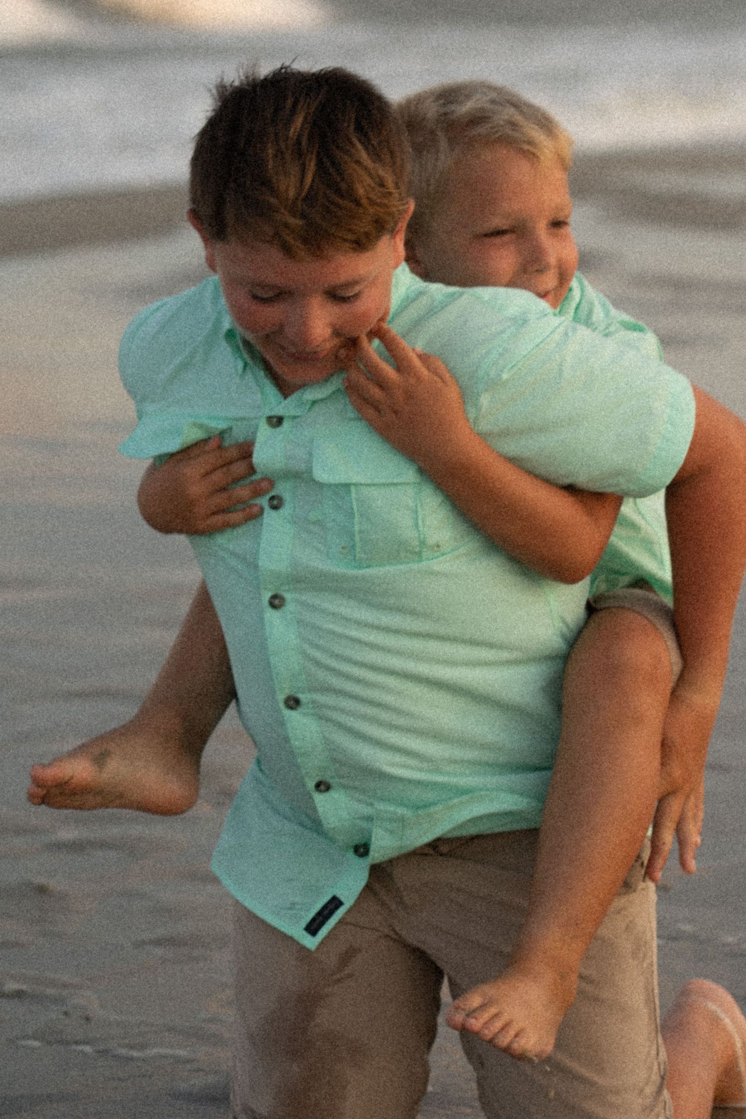 Two boys in matching mint green shirts playing piggyback on a sandy beach at sunset.