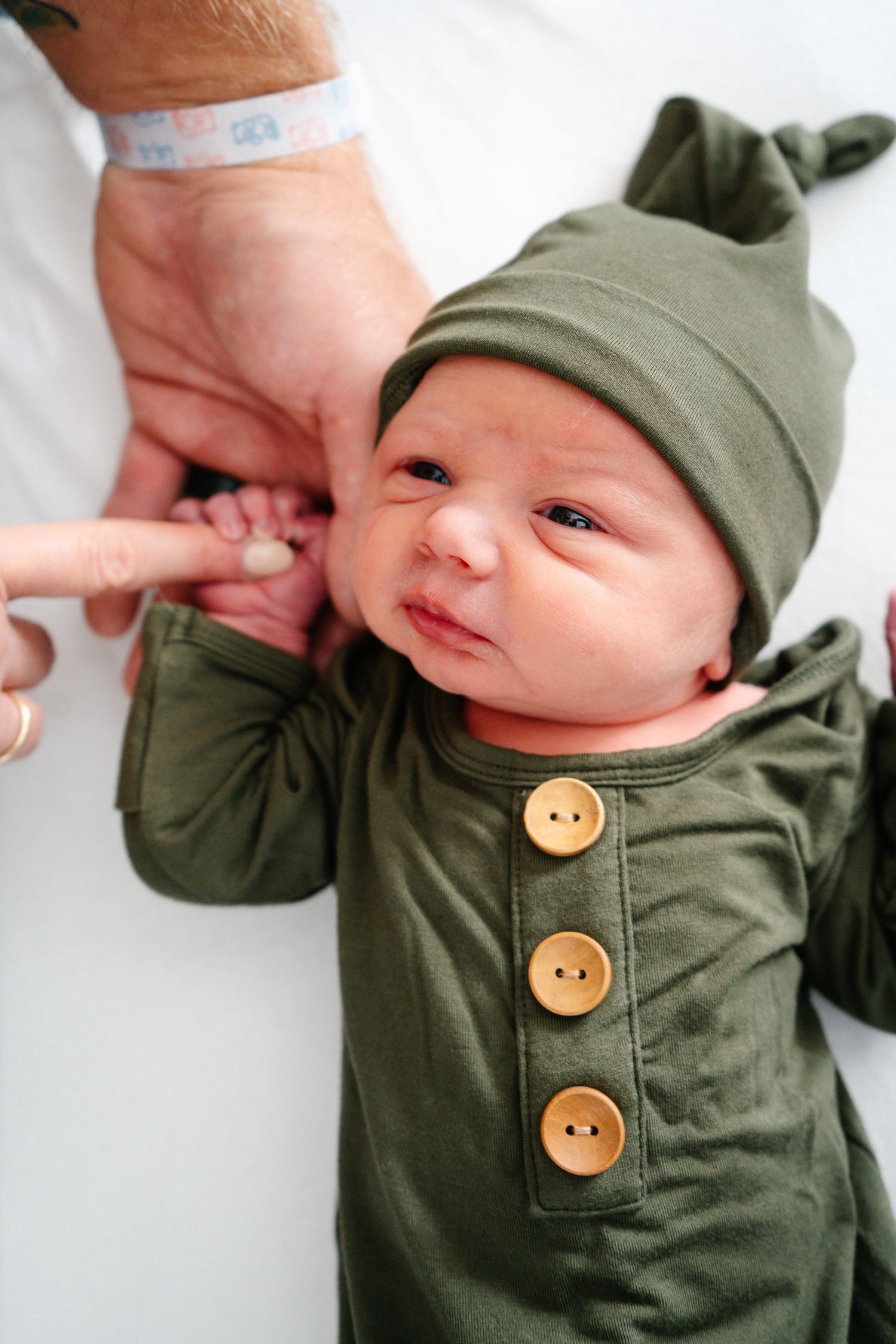 A newborn baby in a green knit outfit and hat grips an adult's finger.