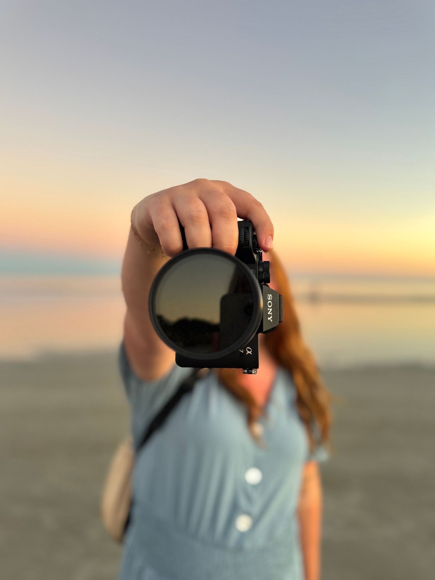 A woman holds a Sony camera toward the lens at a beach during a golden sunset.