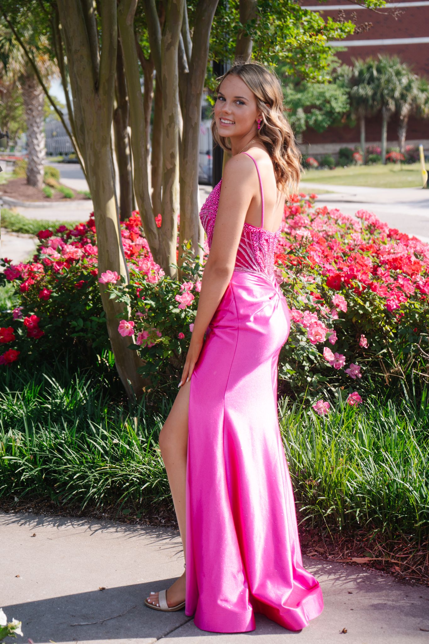 Smiling young woman in hot pink slit formal gown posing beside blooming rose bushes outdoors.