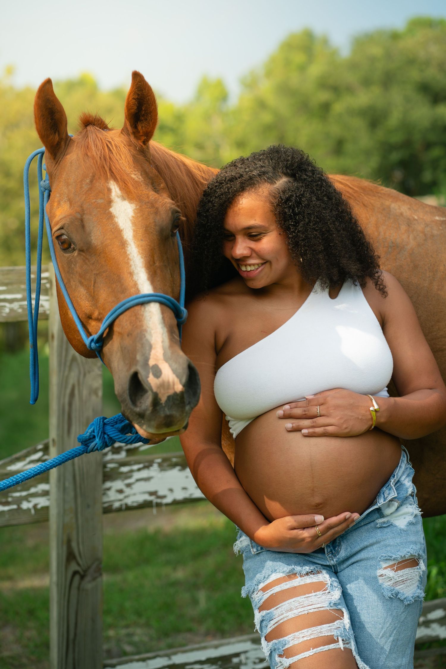 Smiling pregnant woman in white crop top and ripped jeans cradling her baby bump beside a chestnut horse outdoors.