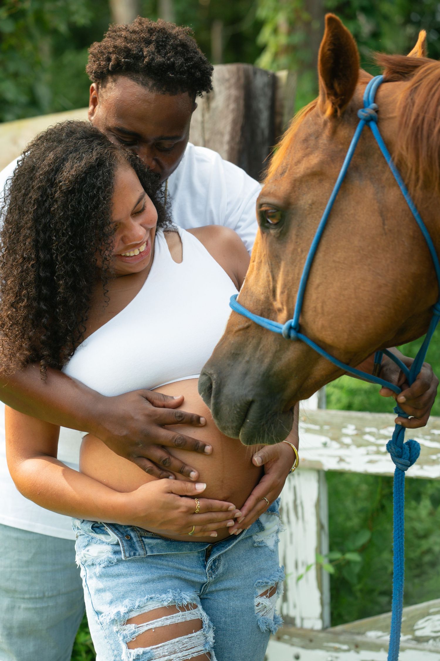 Smiling couple with pregnant belly posing next to a brown horse outdoors.