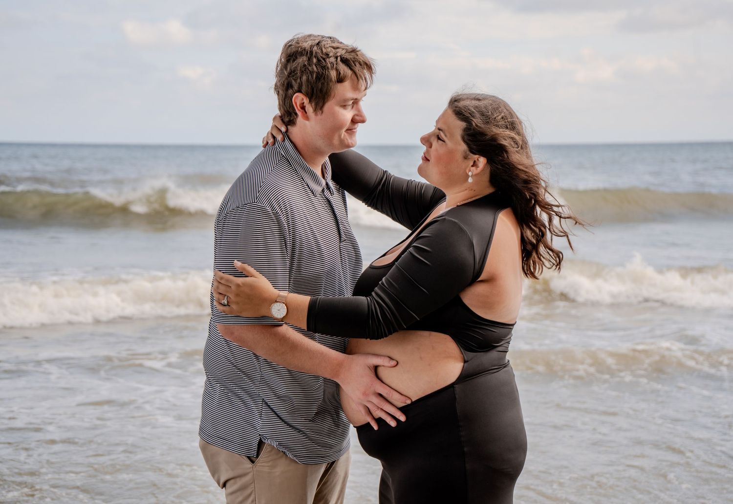 Couple embracing on beach shore with ocean waves, man holding pregnant woman's belly.