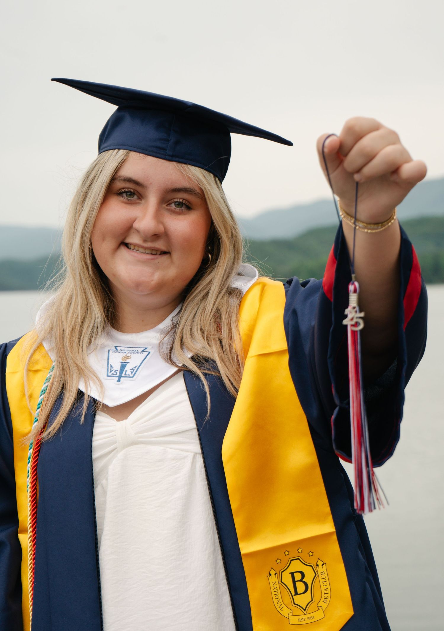 Smiling blonde graduate in navy and gold cap and gown holding tassel with lake backdrop.