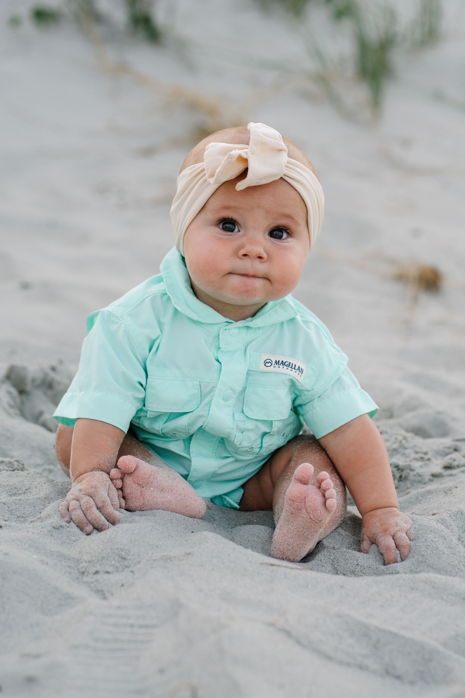 Adorable baby girl in mint green shirt and tan bow headband sitting barefoot on sandy beach.