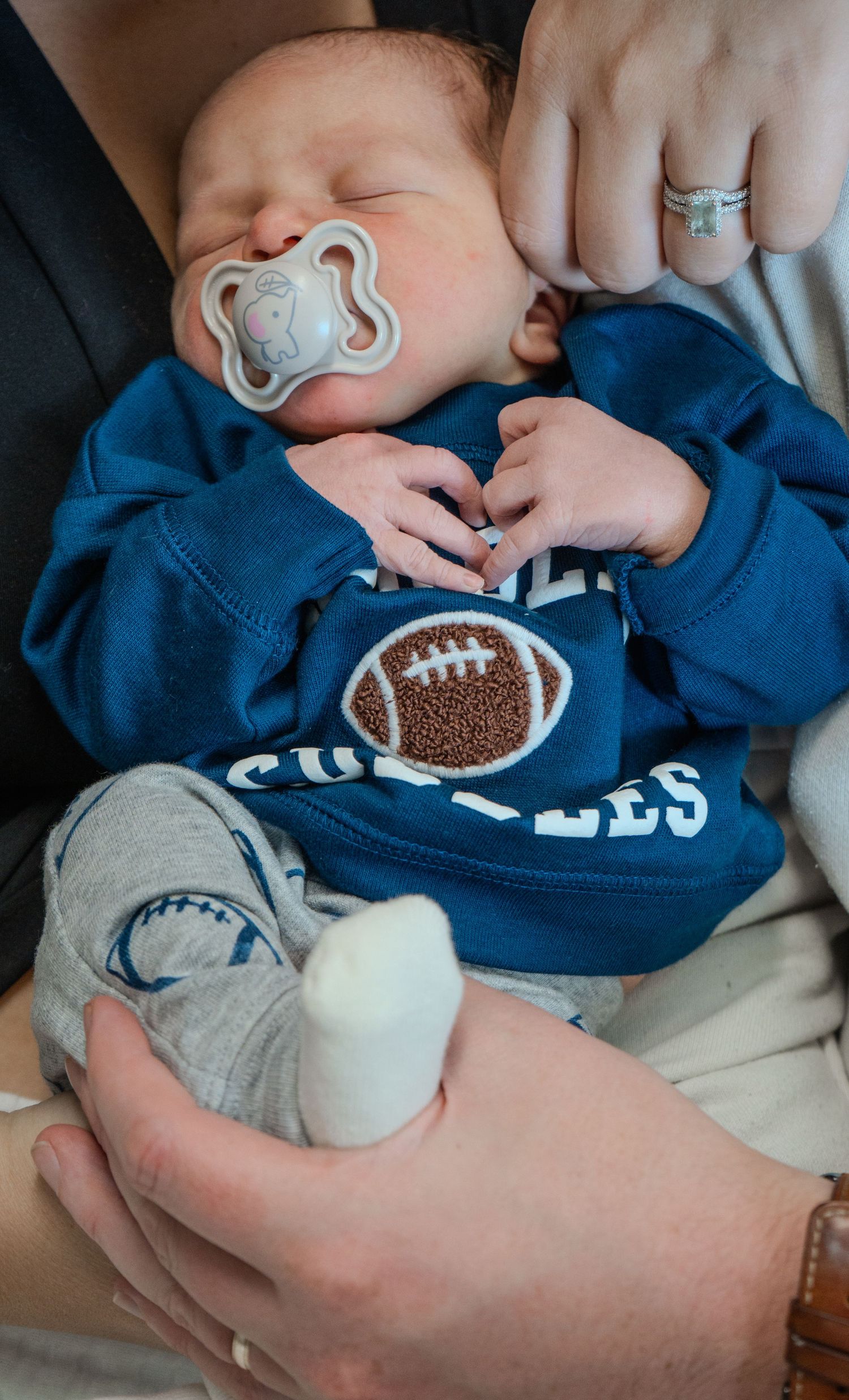 Sleeping newborn baby in blue football onesie with pacifier, cradled in adult hands.