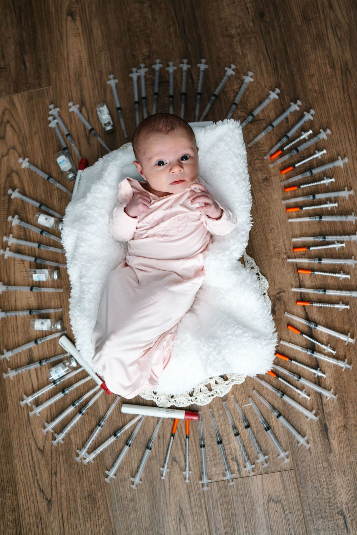 IVF photo, Newborn baby in pink onesie lying on white blanket surrounded by medical syringes arranged in oval pattern on wood floor.