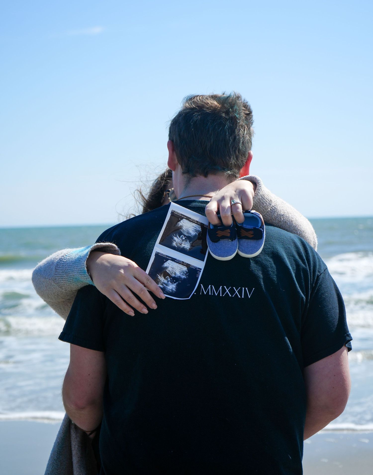 Couple embracing at beach holding ultrasound photos and baby shoes, MMXXIV shirt, pregnancy announcement photo.