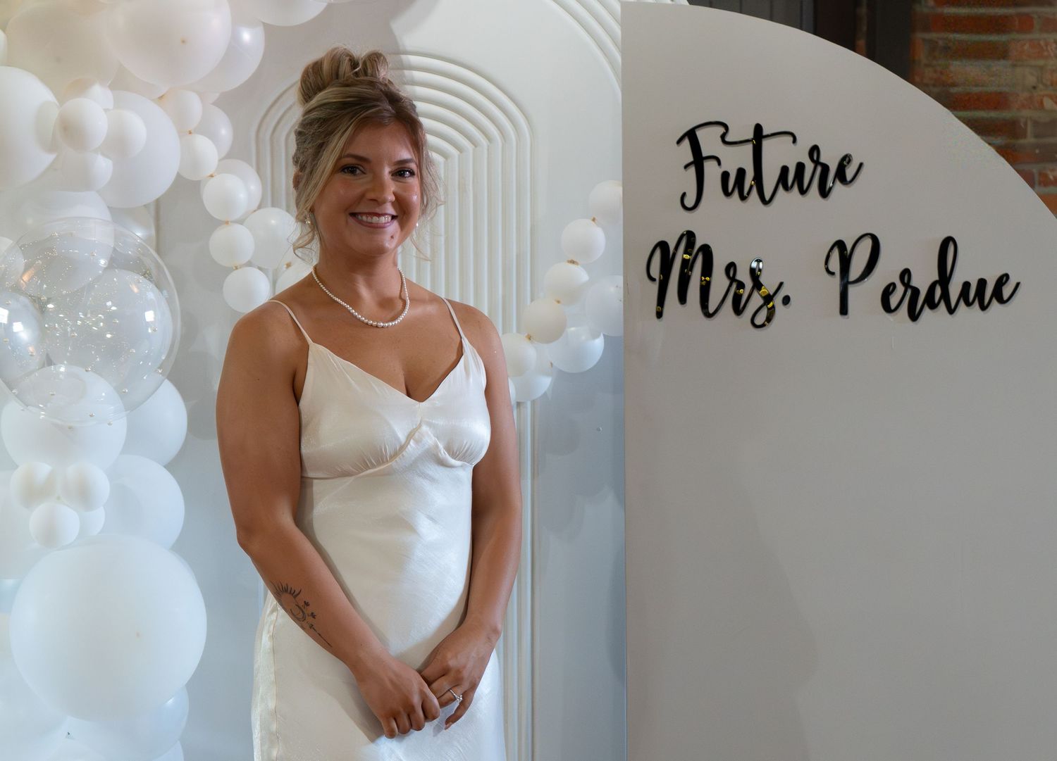 Bride-to-be in white dress smiling beside Future Mrs. Perdue sign at bridal shower with balloon backdrop.