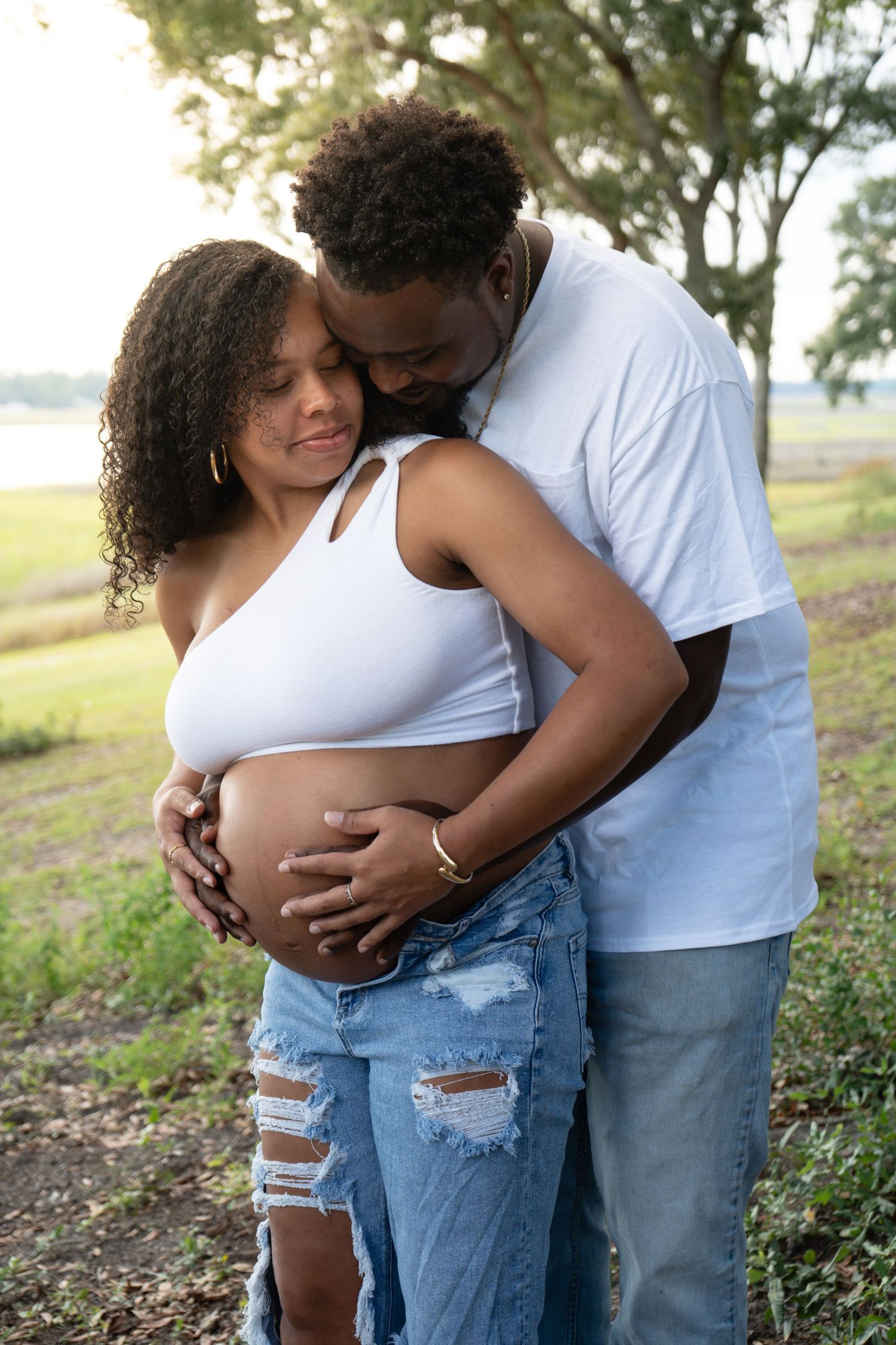 Couple embracing outdoors, man holding pregnant woman's belly near a lake.