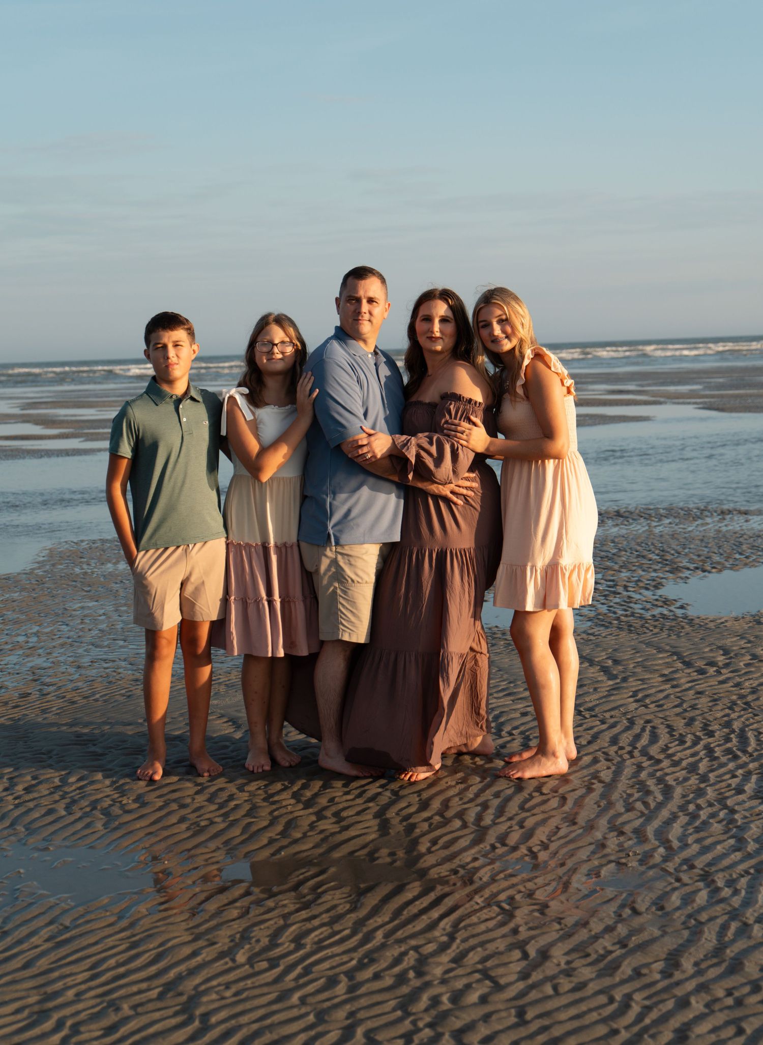 Family of five posing together on sandy beach at golden hour in coordinated casual outfits.