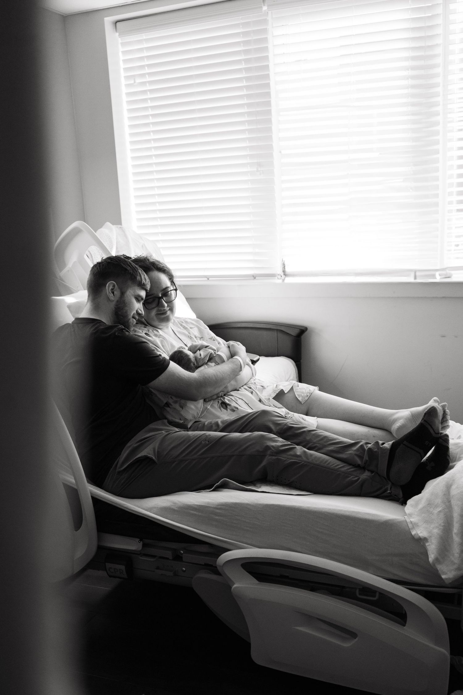 Black and white photo of new parents cuddling newborn baby in hospital bed by window after birth.