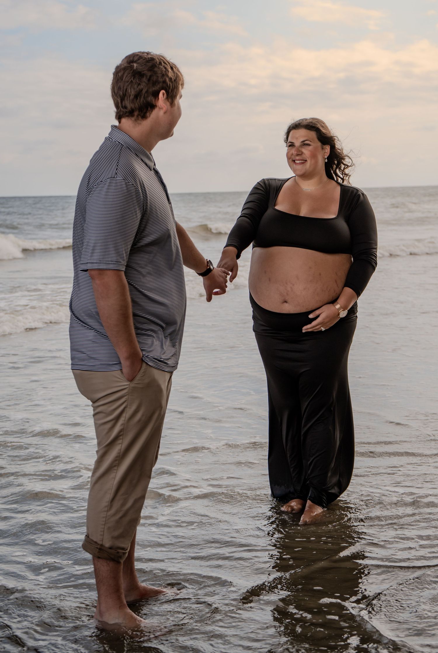 Couple holding hands and smiling at each other while standing in ocean waves.