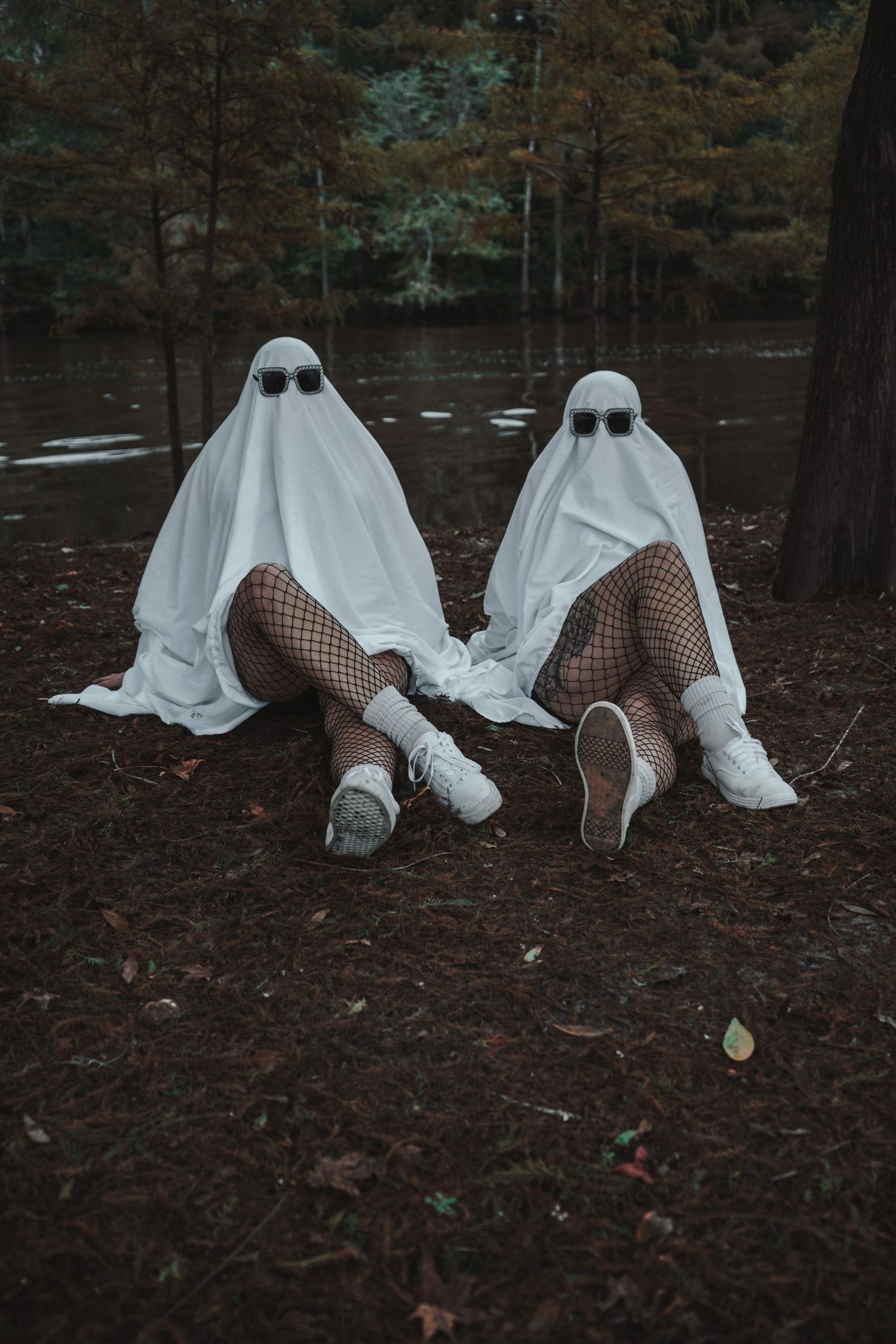 Two people in white ghost Halloween costumes sitting outdoors near a lake.