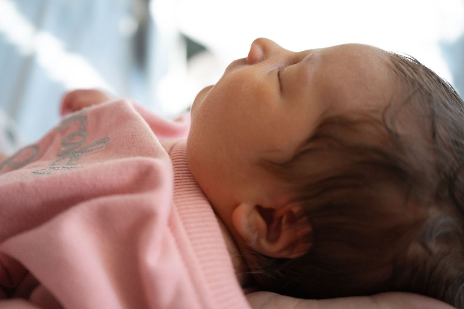 Close-up of a sleeping newborn baby in a pink blanket with dark hair visible against soft light.