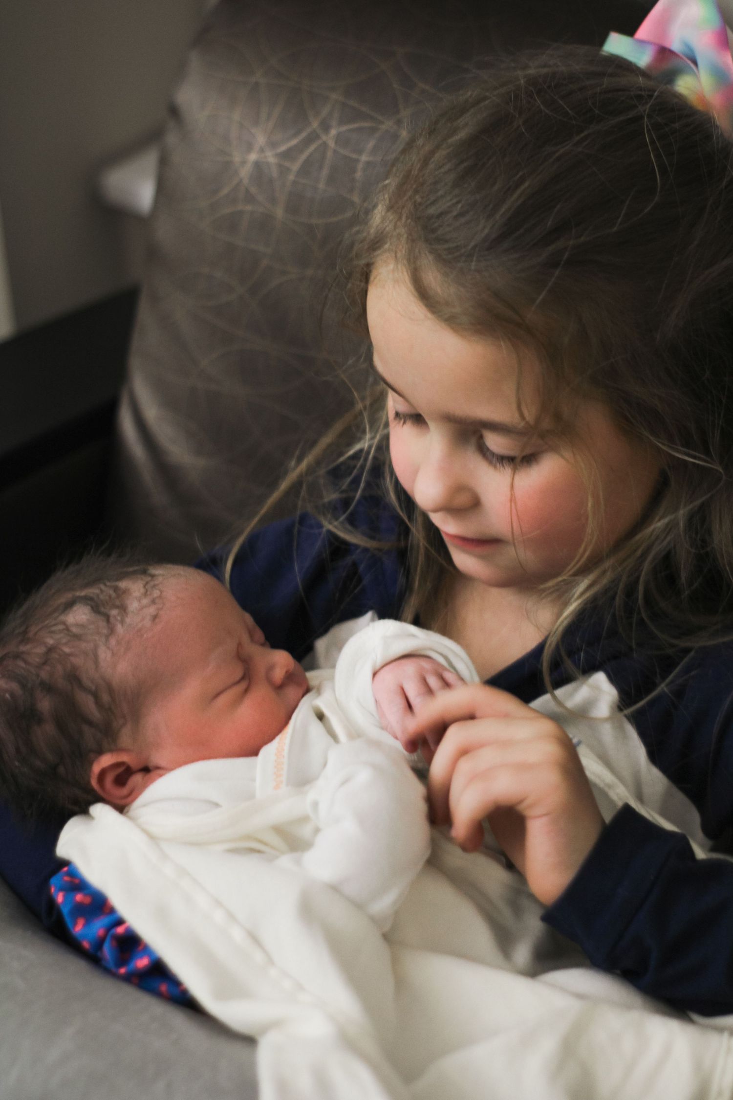 Young girl tenderly holds sleeping newborn baby wrapped in white blanket while seated in gray chair.