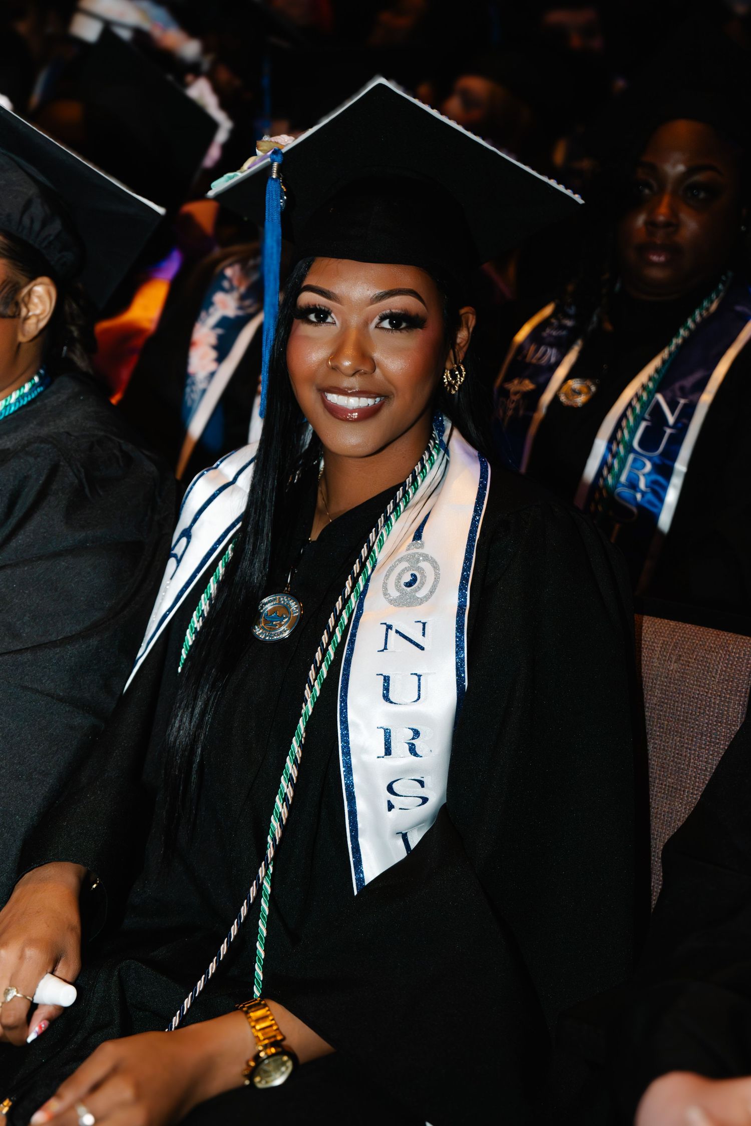 Smiling nursing graduate in black cap and gown wearing a Nursing sash and green honor cords at graduation ceremony.