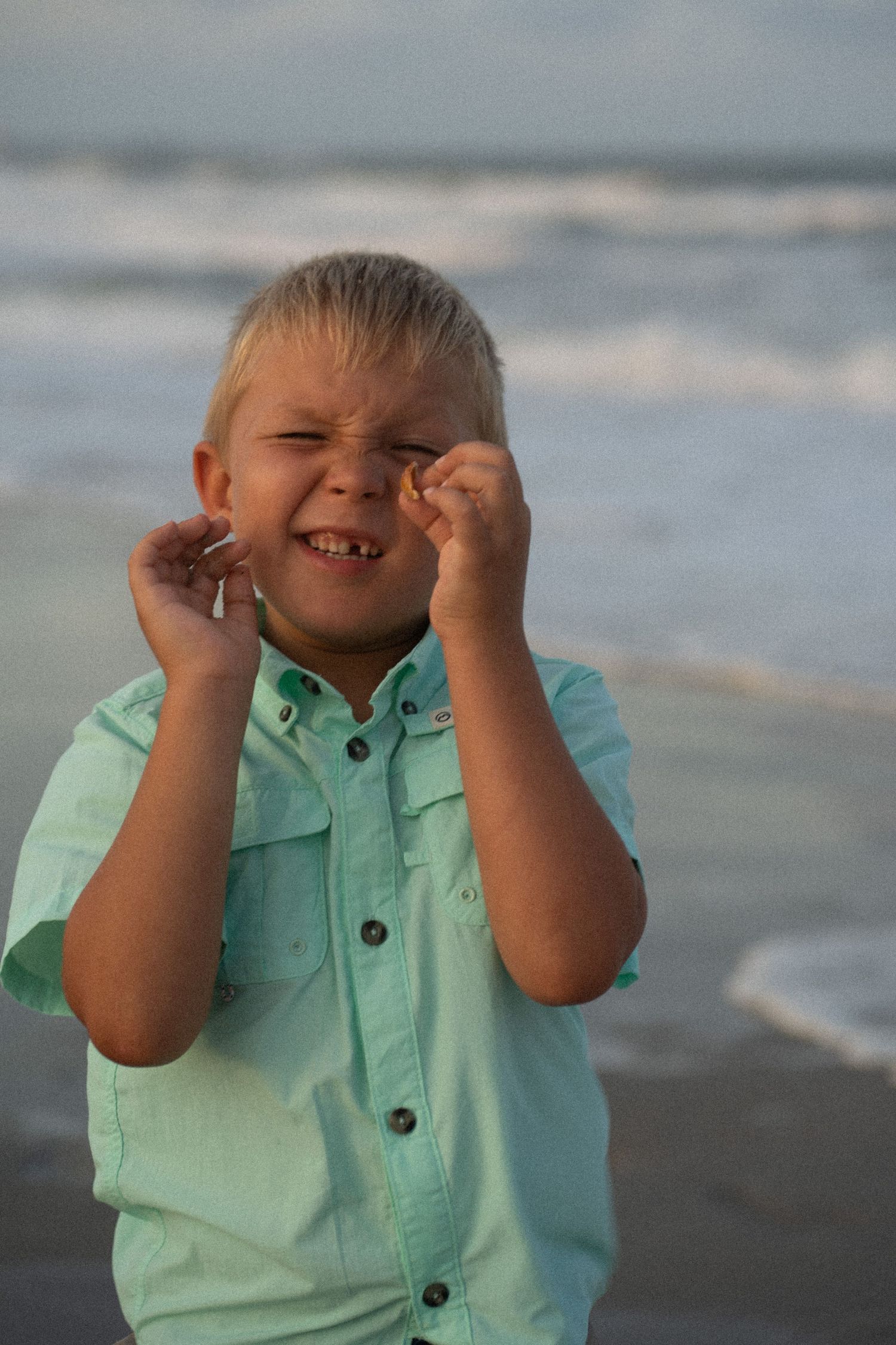 A smiling blonde boy in a mint green shirt poses playfully on a beach with ocean waves in the background.