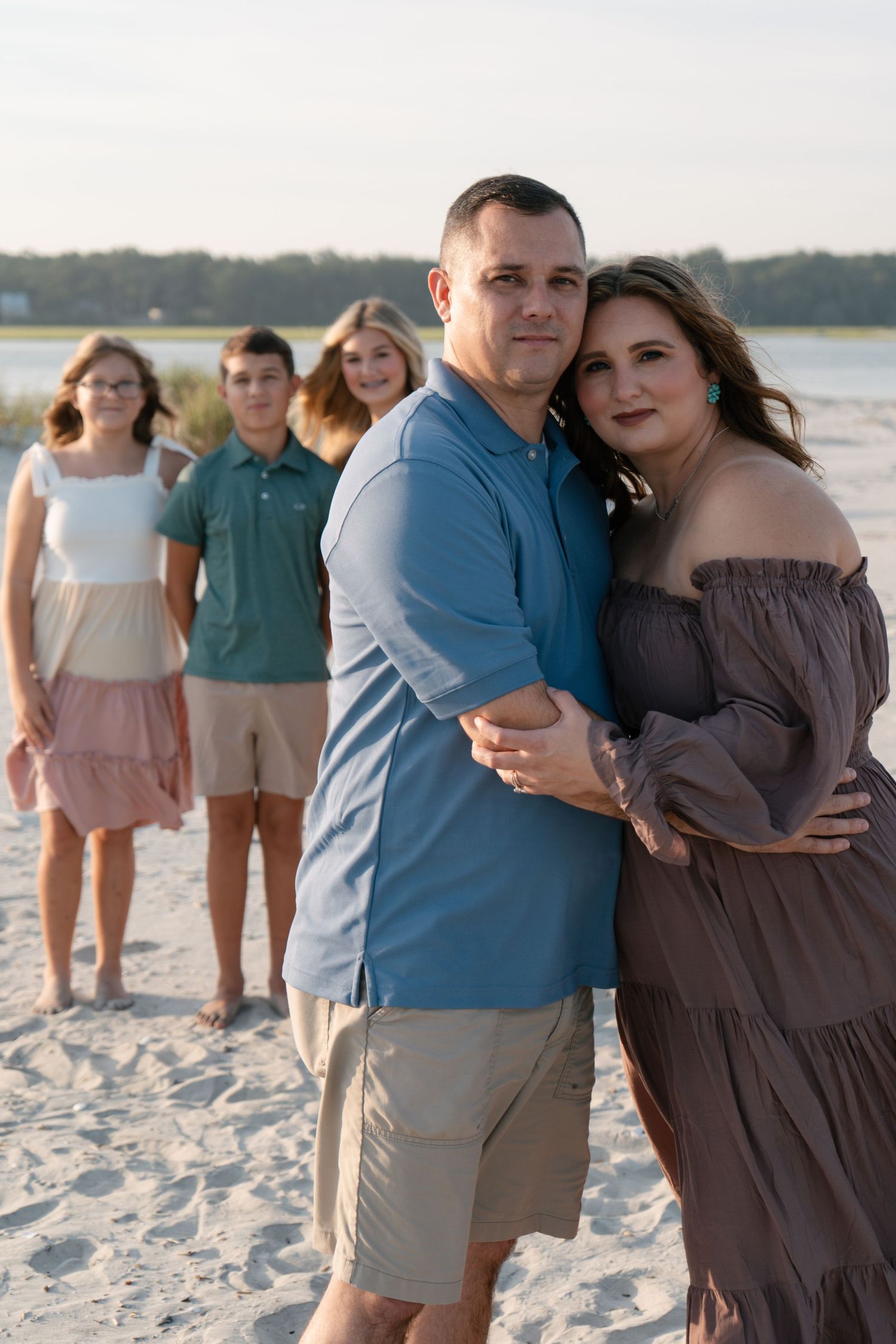 Couple embracing on a beach with their four children standing behind them at sunset.