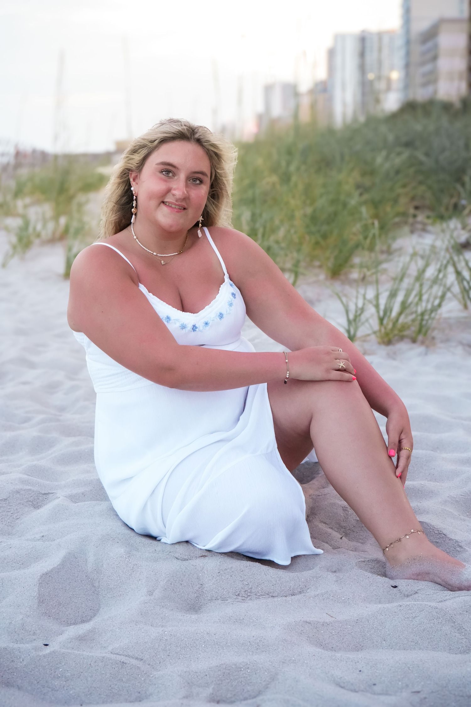Smiling blonde woman in white sundress sitting on sandy beach with ocean and buildings in background.