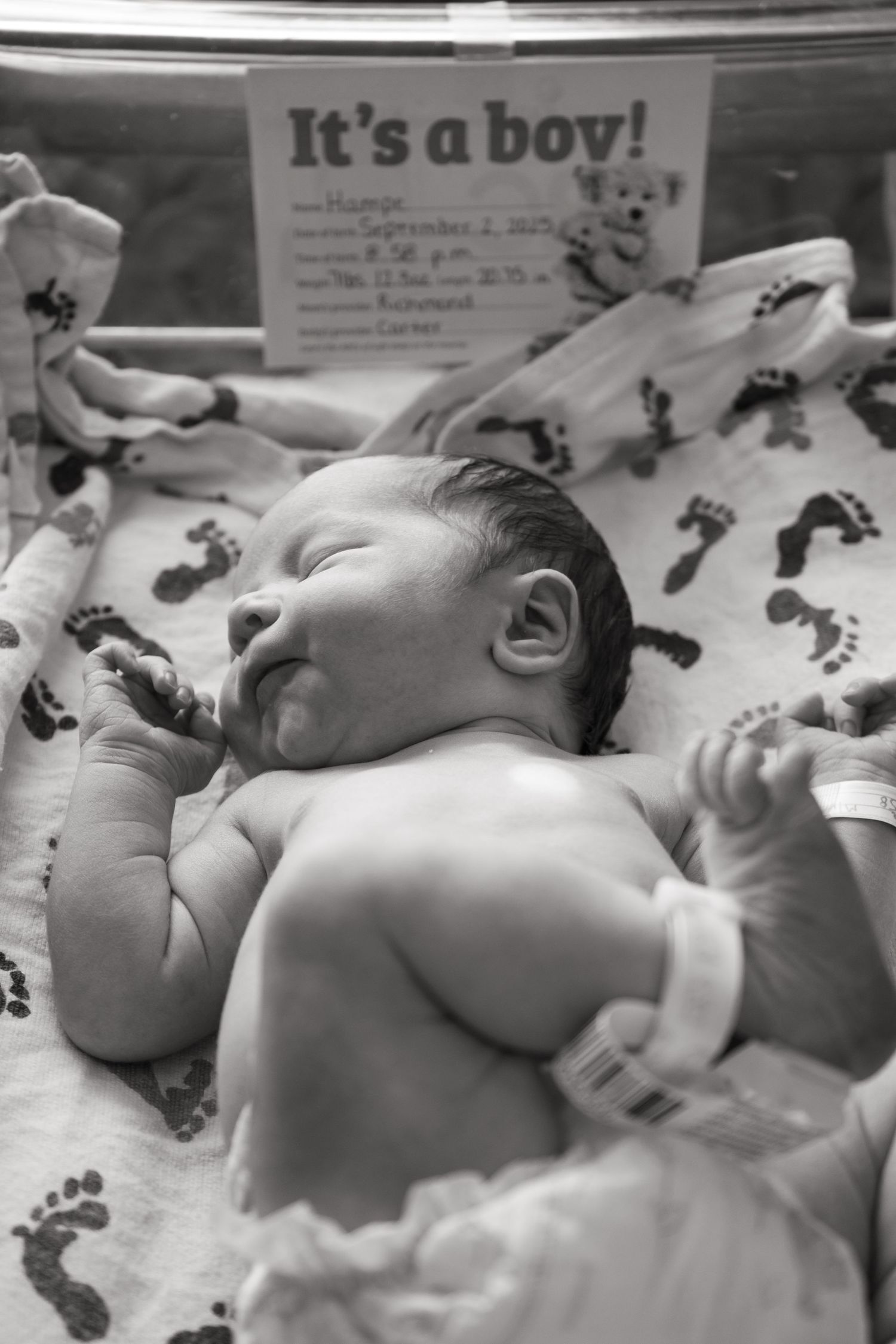 Black and white photo of a newborn baby boy lying in hospital bassinet with It's a Boy sign.