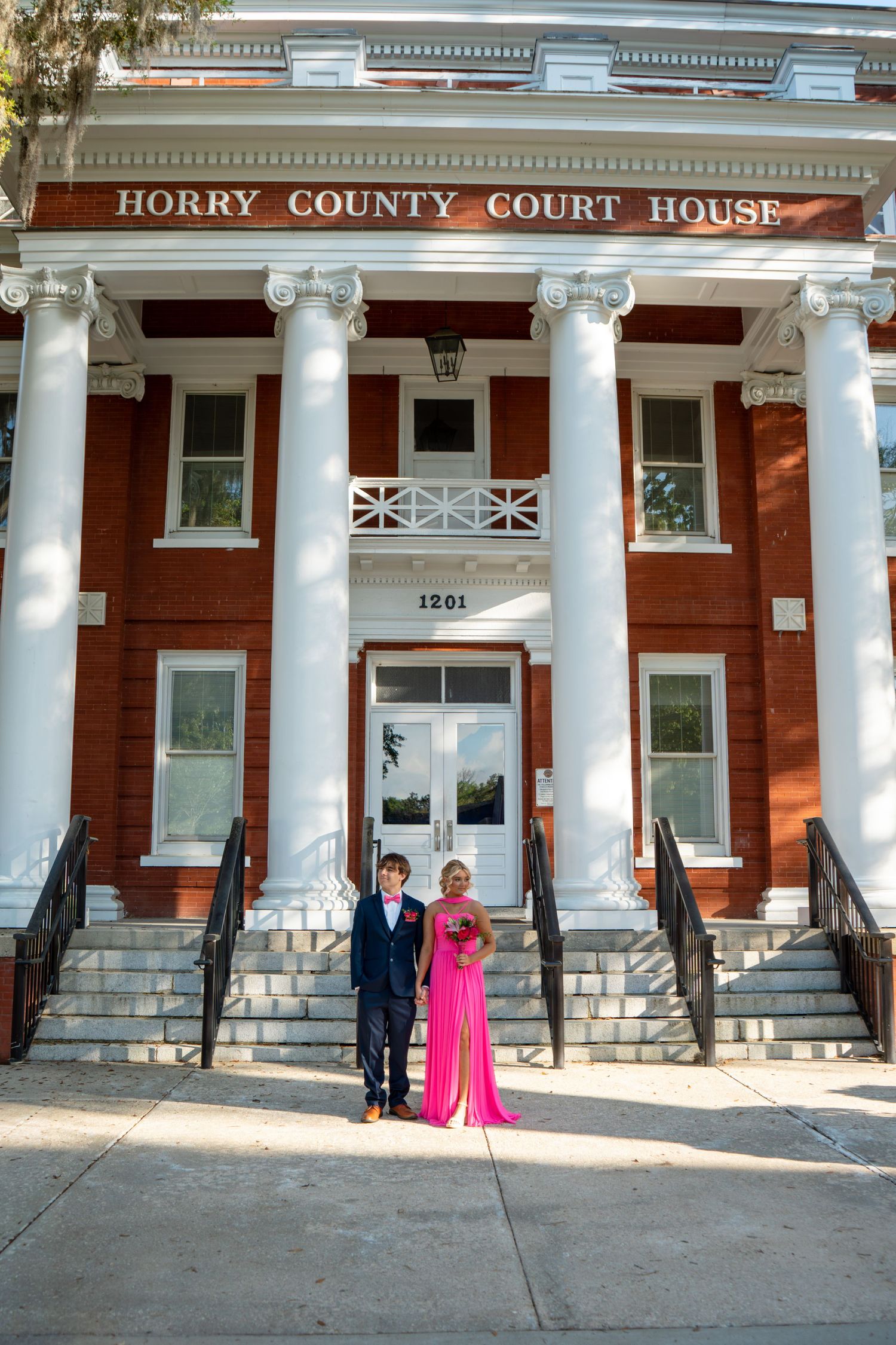 Prom couple posing in front of Horry County Court House, girl in pink gown and boy in blue suit.
