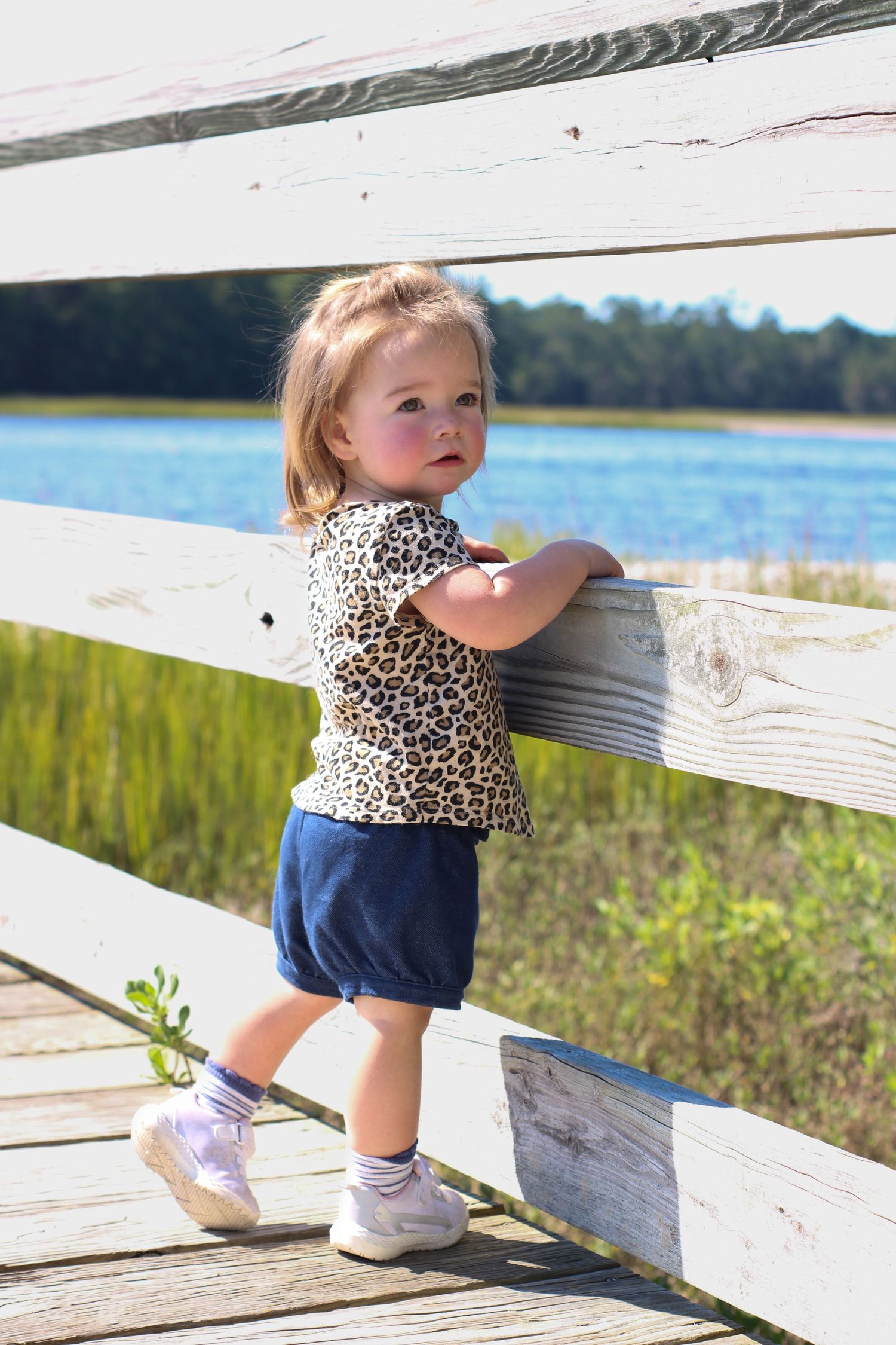 Adorable toddler in leopard print top and navy shorts standing by white wooden fence near lake.