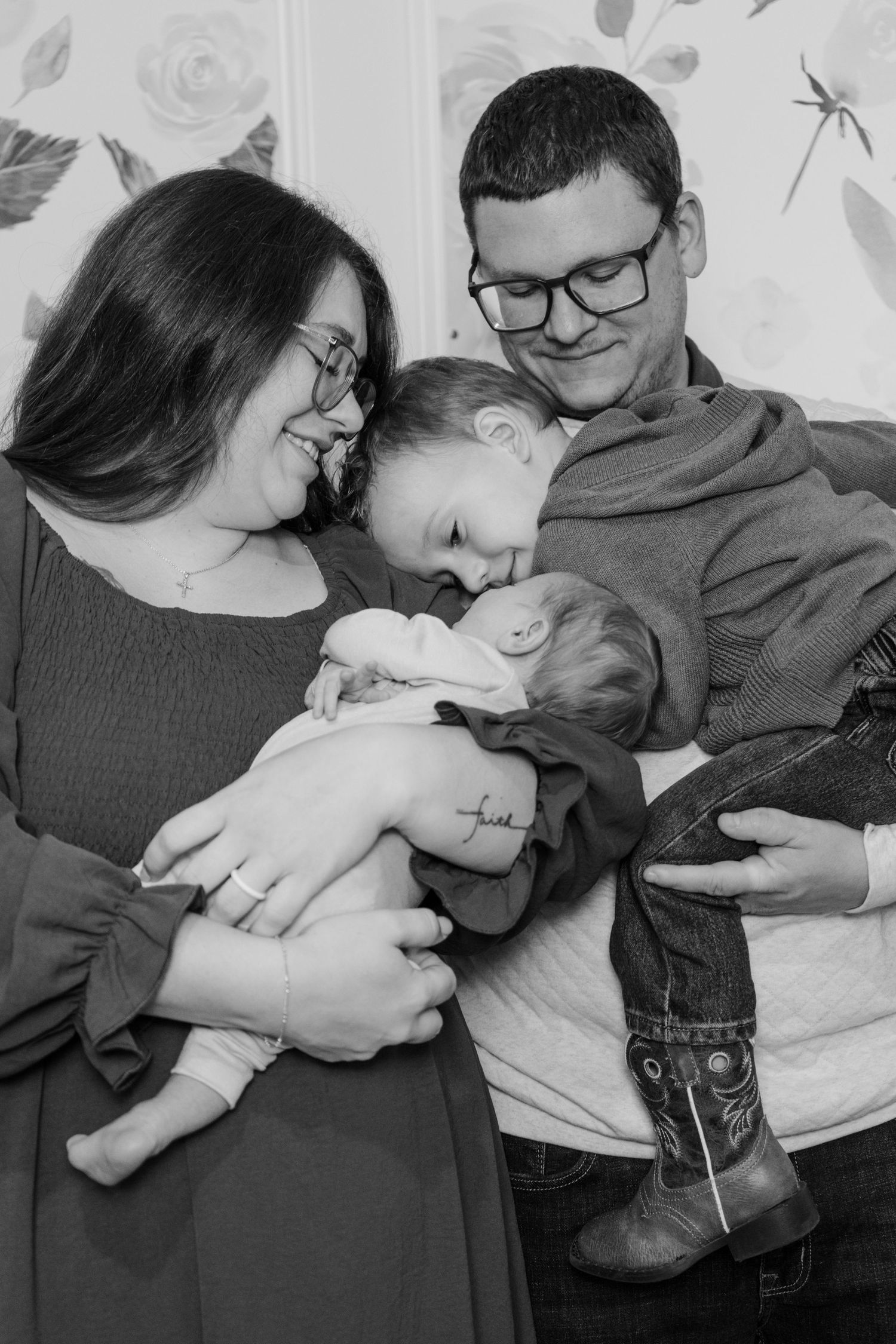 Black and white family portrait of parents with glasses holding a newborn baby and toddler, smiling warmly indoors.
