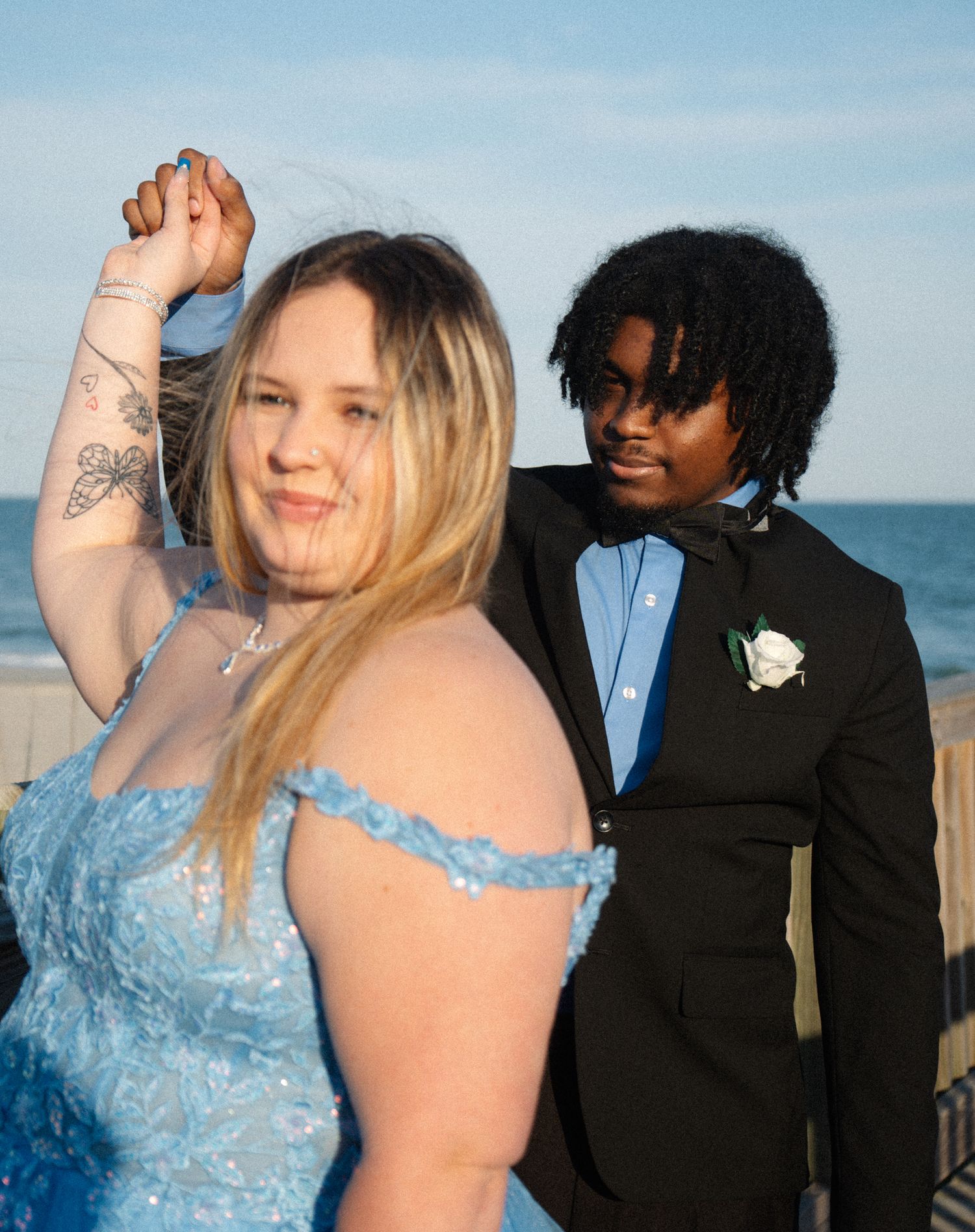 Prom couple in blue dress and black tuxedo dancing outdoors near ocean at sunset.